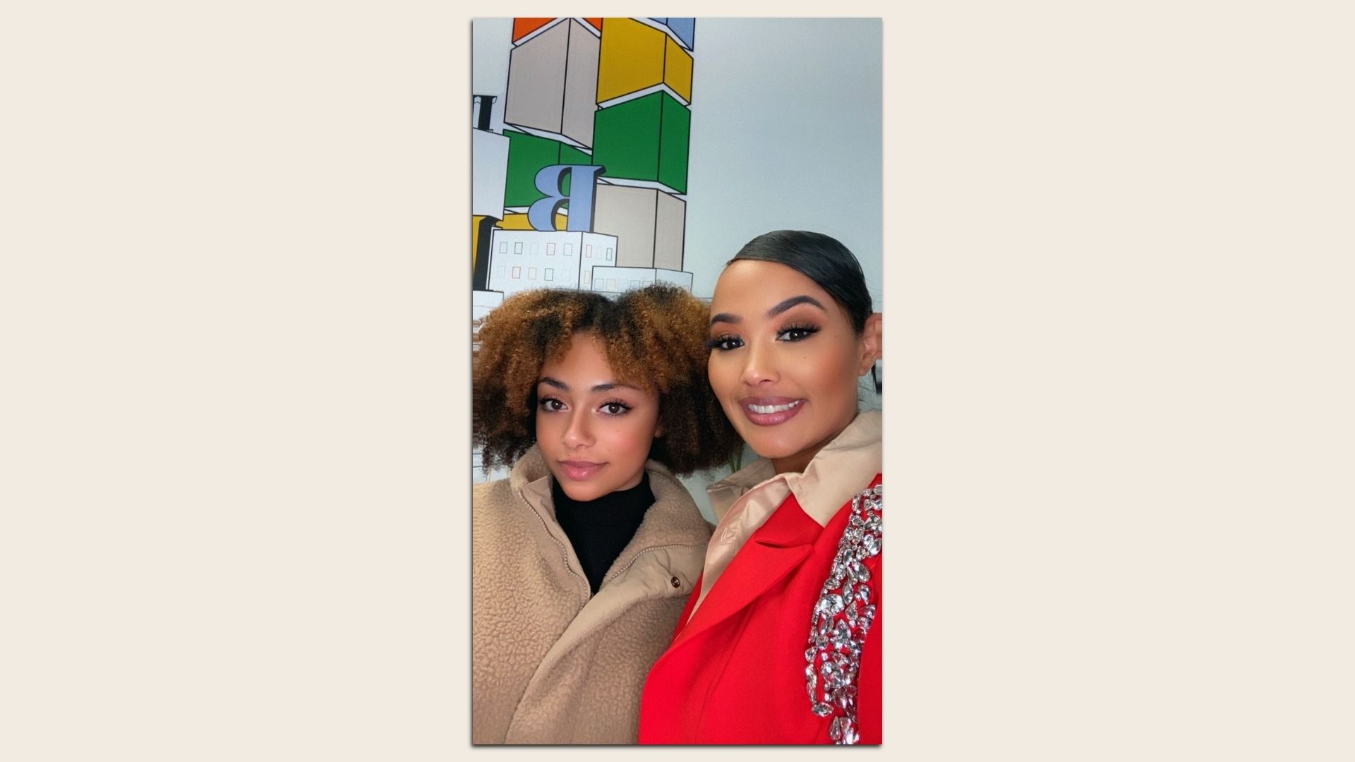 Two women pose for a selfie indoors; the left has curly hair in a beige coat, the right in a red outfit with rhinestones, with colorful stacked-block artwork behind them.