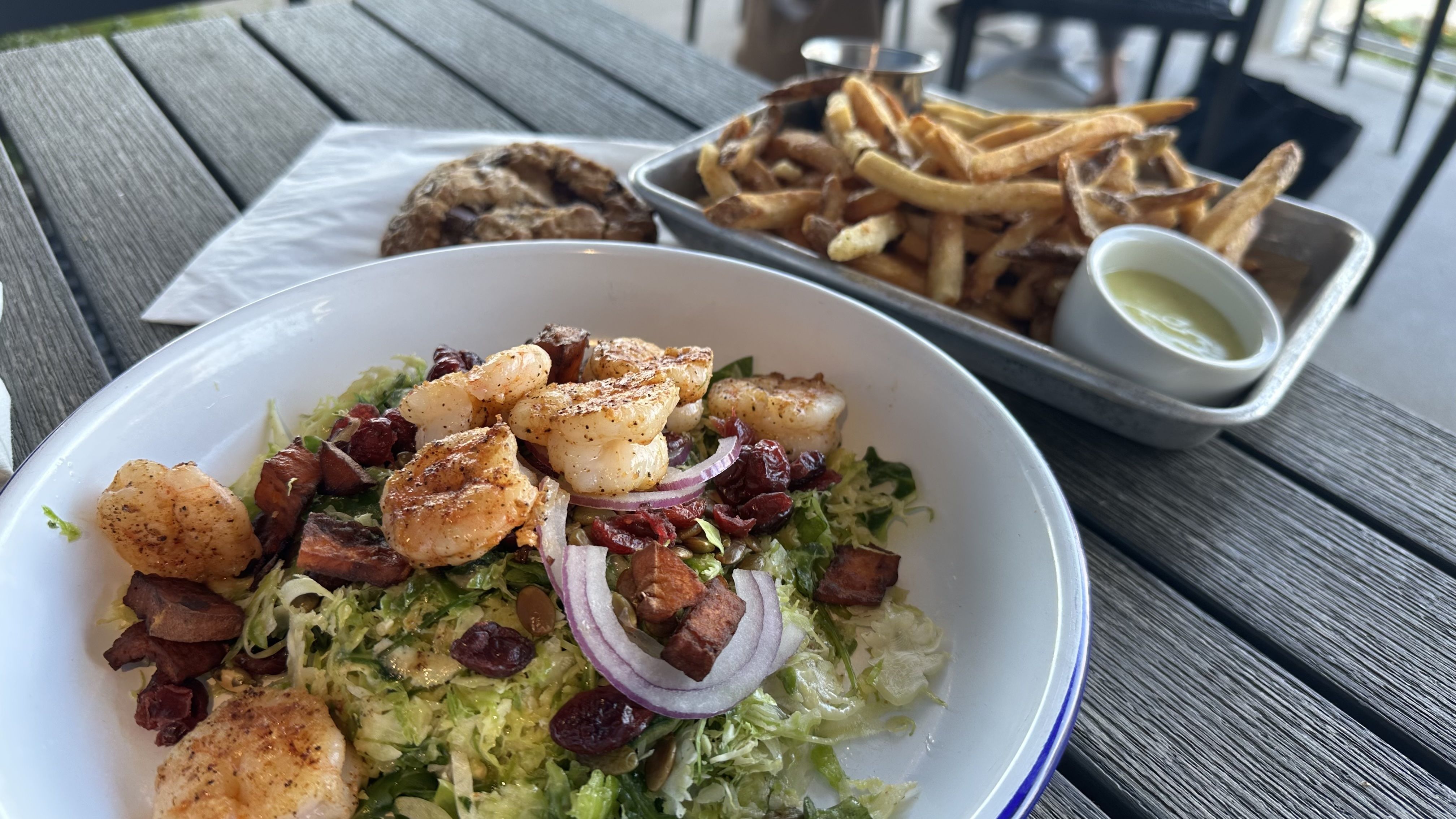 Grilled shrimp salad with red onions, dried cranberries, and roasted sweet potato in a white bowl. Fries and dipping sauce in a metal tray, with a chocolate chip cookie on a piece of paper nearby.