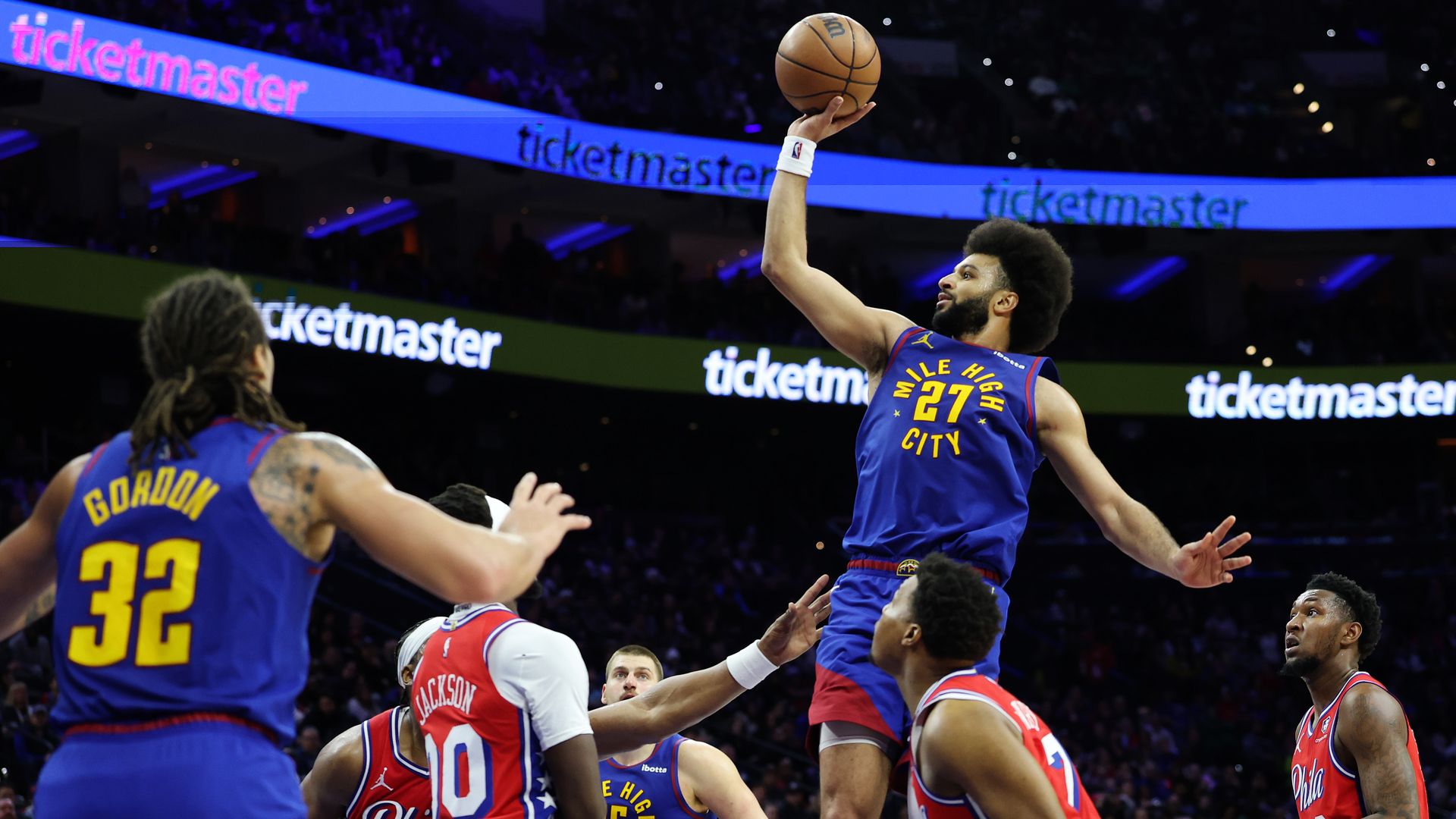 A man in a blue basketball jersey with the word MILE HIGH CITY and number twenty-seven rises over other players with a ball in his hand. 