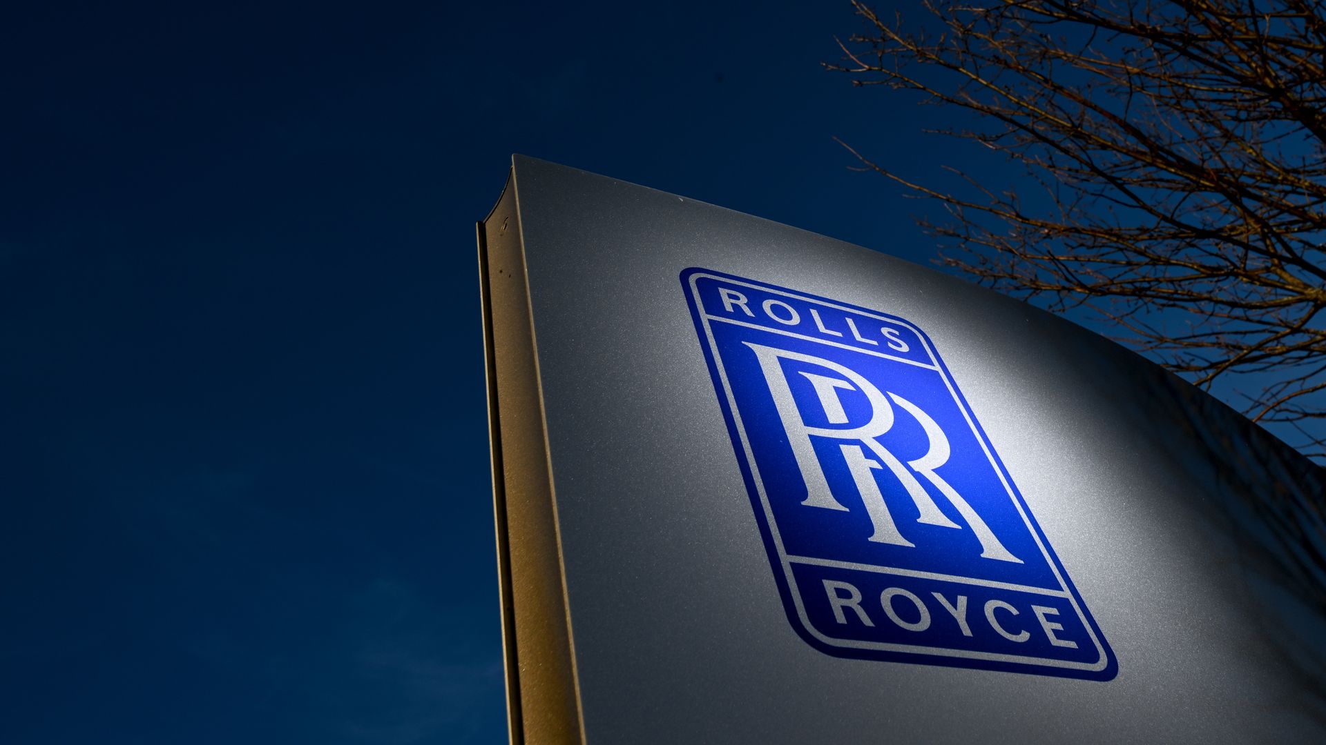 A sign showing the Rolls-Royce logo. The sky behind it is blue. There is a tree on the right side.