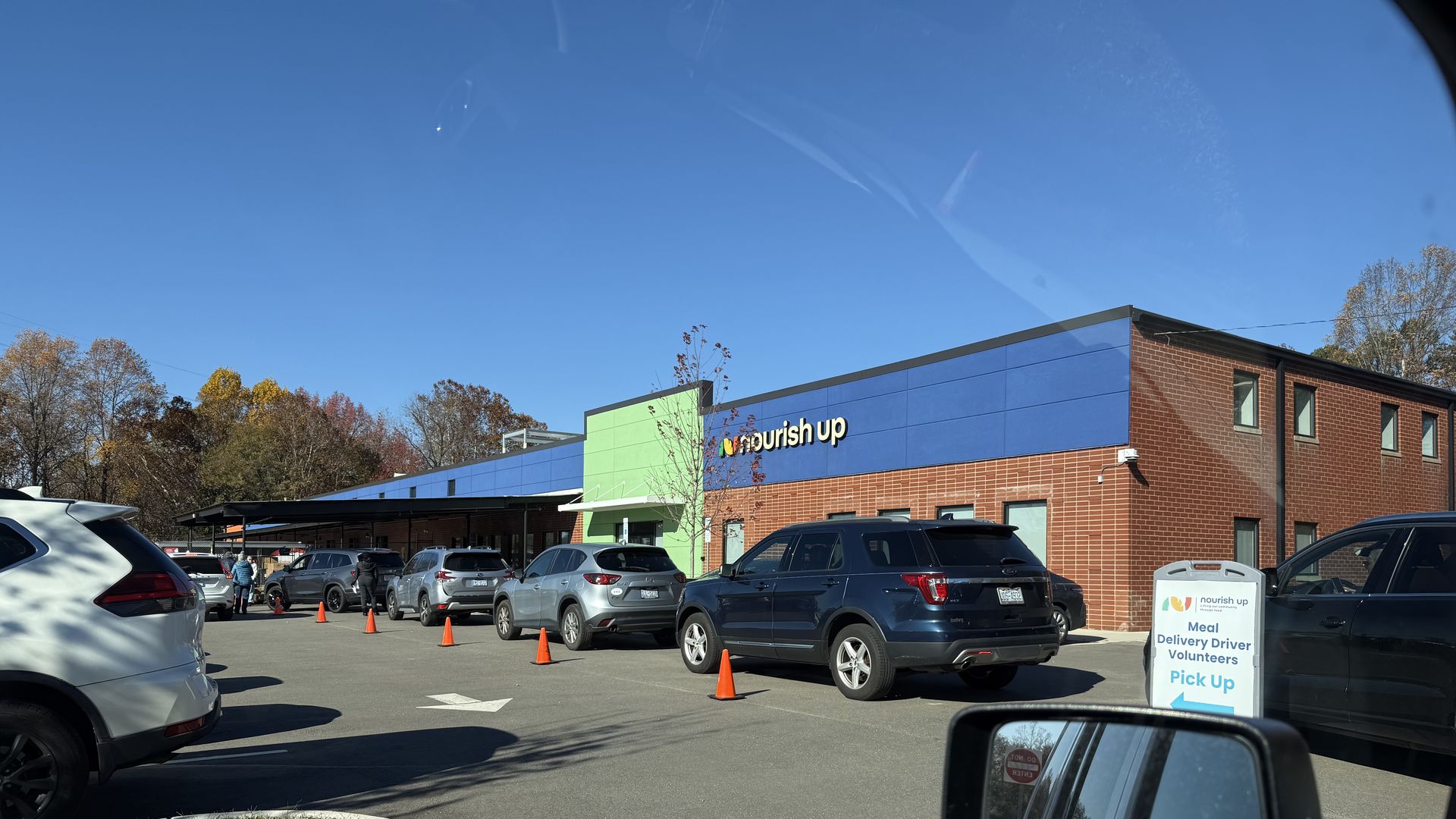 Line of cars and volunteers outside a brick building with blue and green accents at "nourish up" for Meal Delivery Driver Volunteer Pick Up on a clear, sunny day.