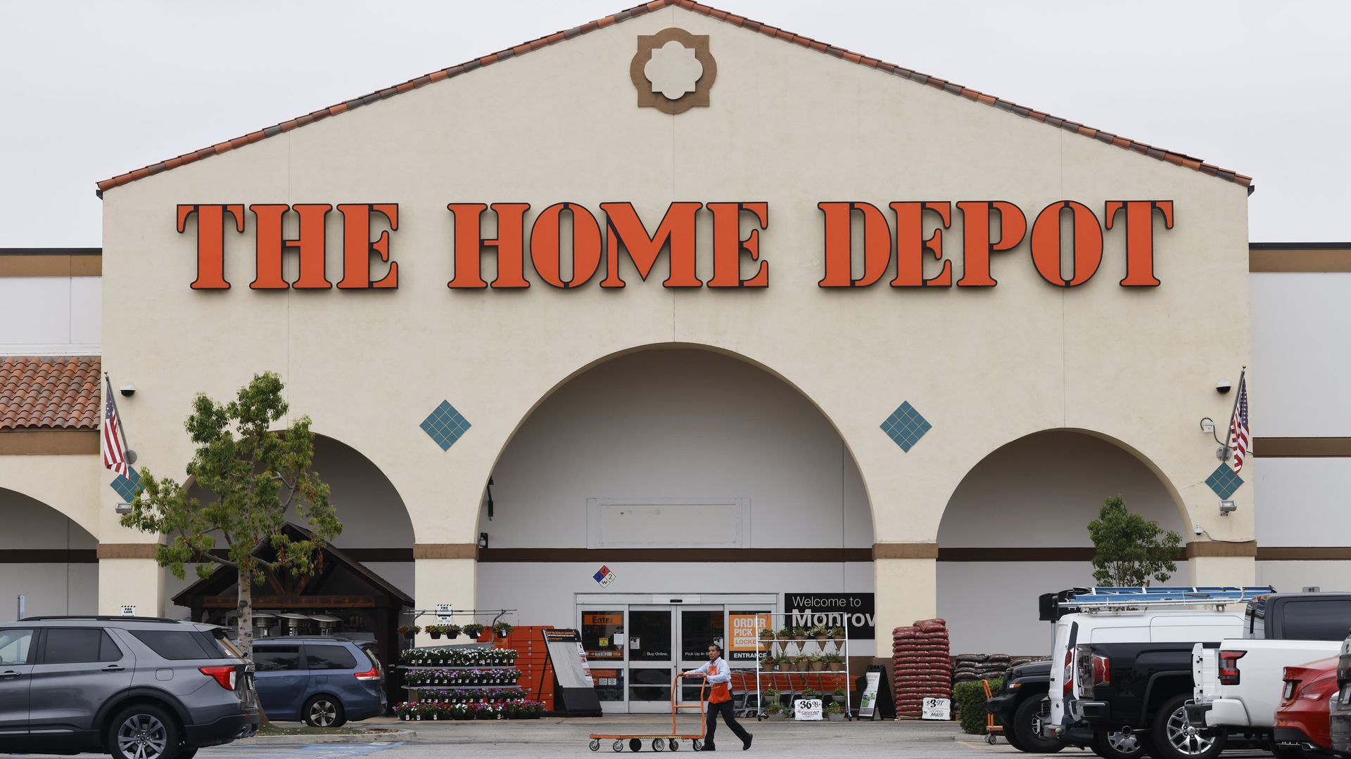 Exterior of The Home Depot store with large orange sign, plants near entrance, an employee pushing a cart, parked cars, and two American flags on a cloudy day.