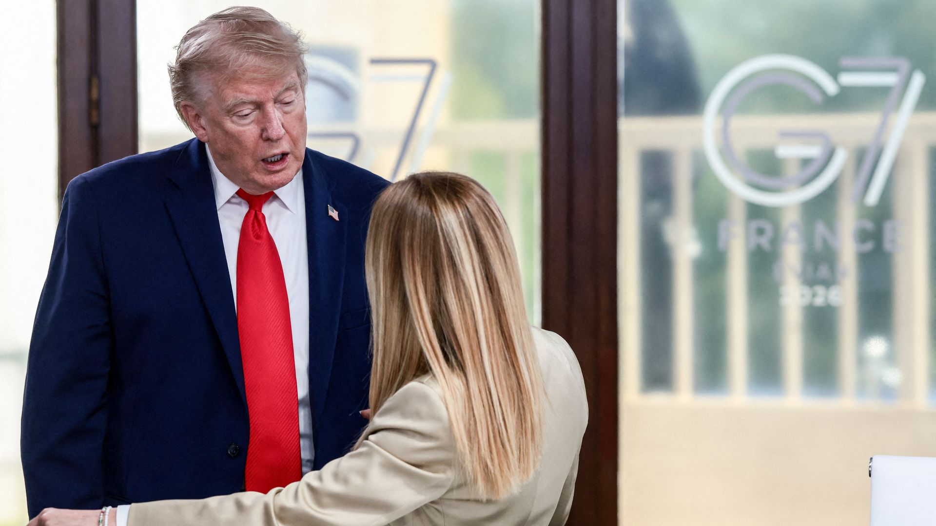 President Trump, in a suit and red tie, speaks to a woman with her back to the camera in front of a "G7" logo.