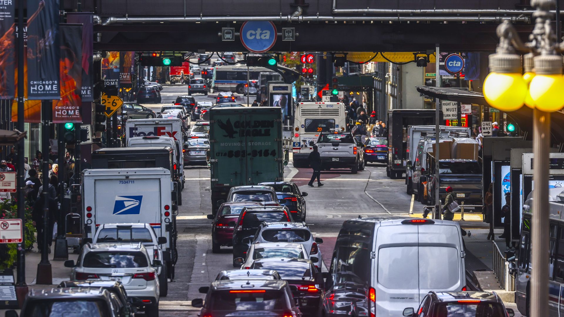 Busy downtown street beneath an elevated rail, with a blue CTA circle sign overhead. Congested traffic includes cars, buses, and a FedEx truck, with pedestrians and banners along the sidewalk.