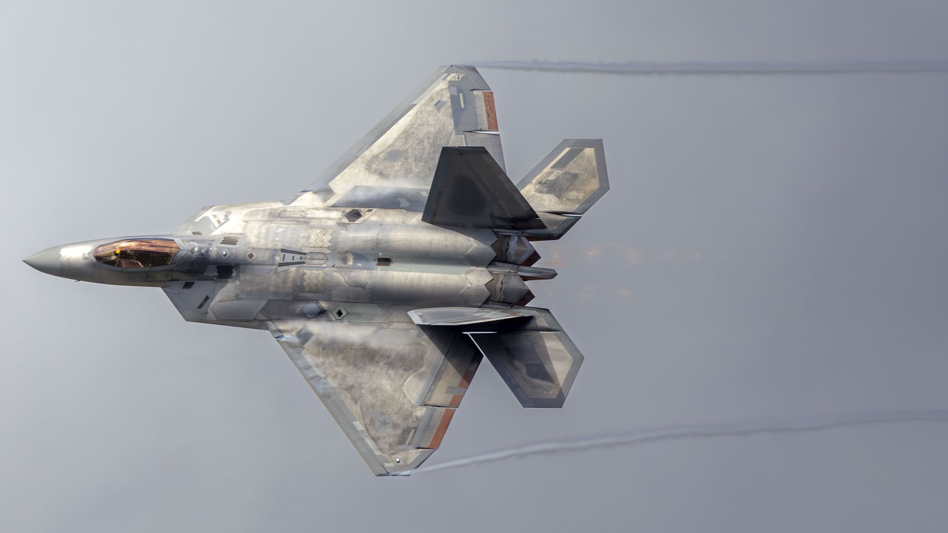 Gray camouflaged F-22 fighter jet flying with afterburner on, leaving faint vapor trails against a clear sky.