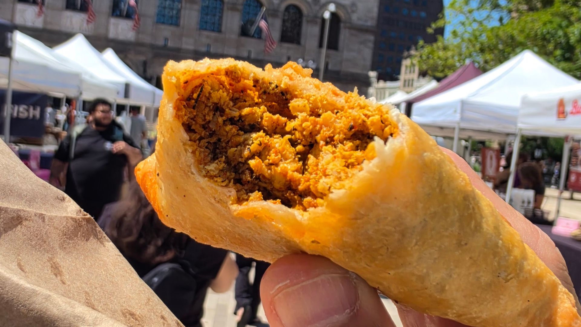 Close-up of a flaky, golden fried pastry filled with orange, crumbly stuffing, held up in a sunny outdoor market with white tents and blurred shoppers in the background.