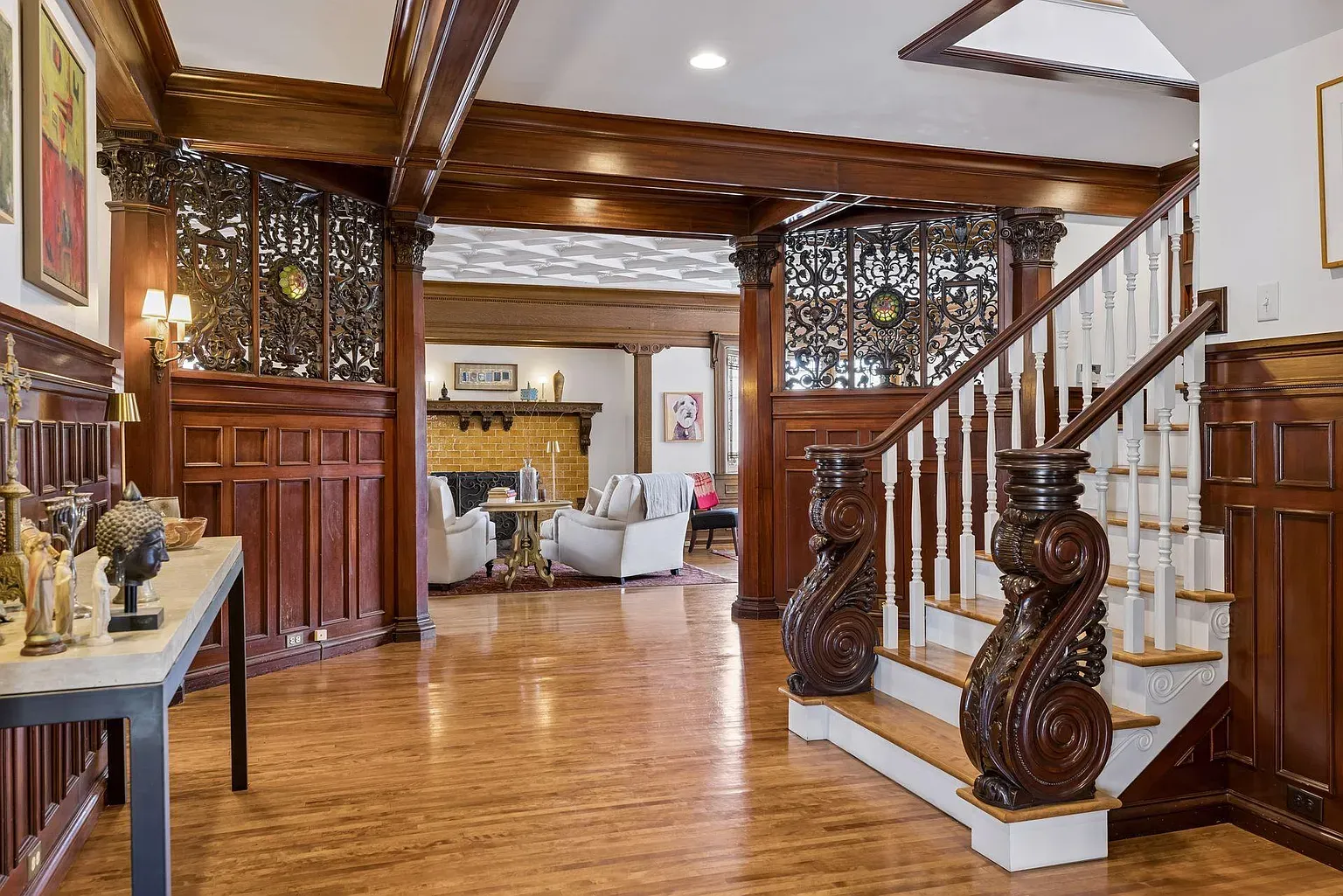 Luxurious foyer with dark wood paneling, ornate wrought-iron screens, and a carved staircase. Warm hardwood floors flow into a sitting area with a brick fireplace and cream seating.
