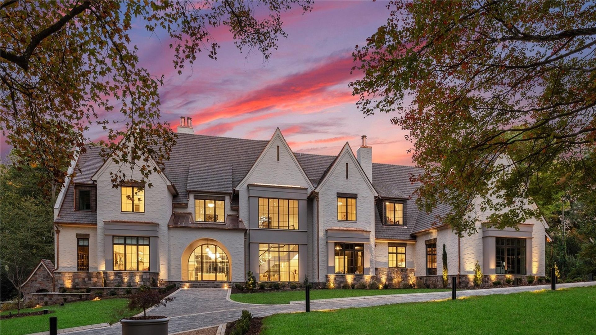 Large white brick house with multiple gables and lit windows at sunset, surrounded by grass and trees with a curved stone driveway and outdoor lighting.