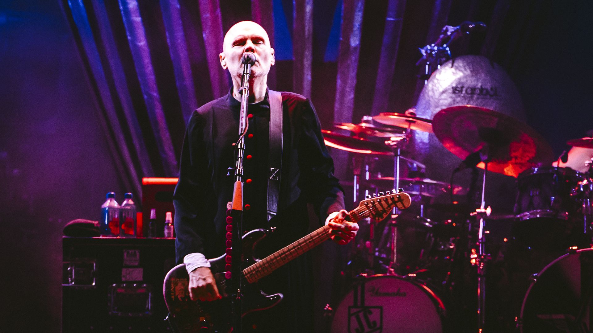 Bald male guitarist and vocalist on stage, dressed in a black coat with red buttons, playing an electric guitar and singing into a microphone. Purple lighting, drum kit visible in the background.
