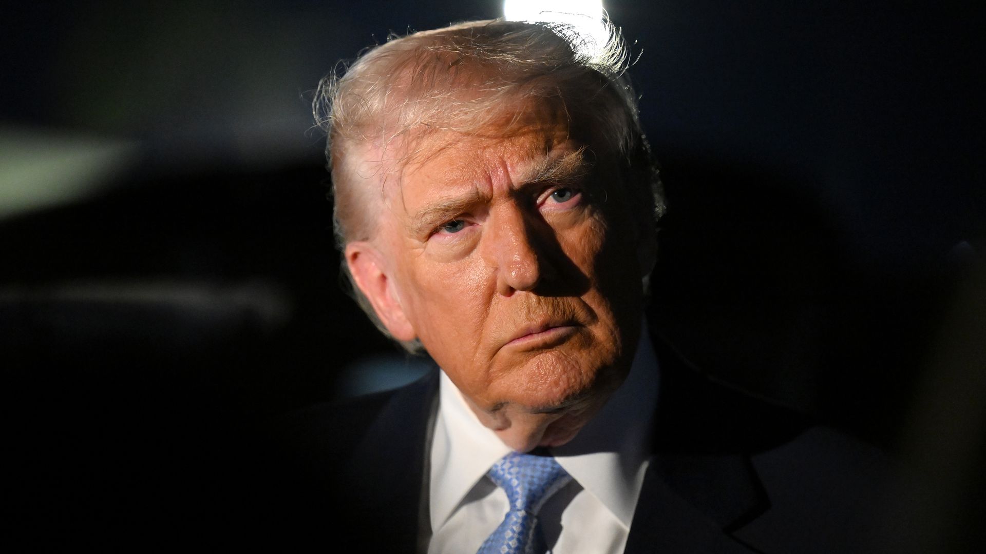 President Trump, wearing a navy jacket with a US flag pin at the top of his left lapel, white shirt and sky-blue tie, speaks to reporters in the dark before boarding Air Force One on November 16, 2025 at Palm Beach International Airport in West Palm Beach, Florida. 