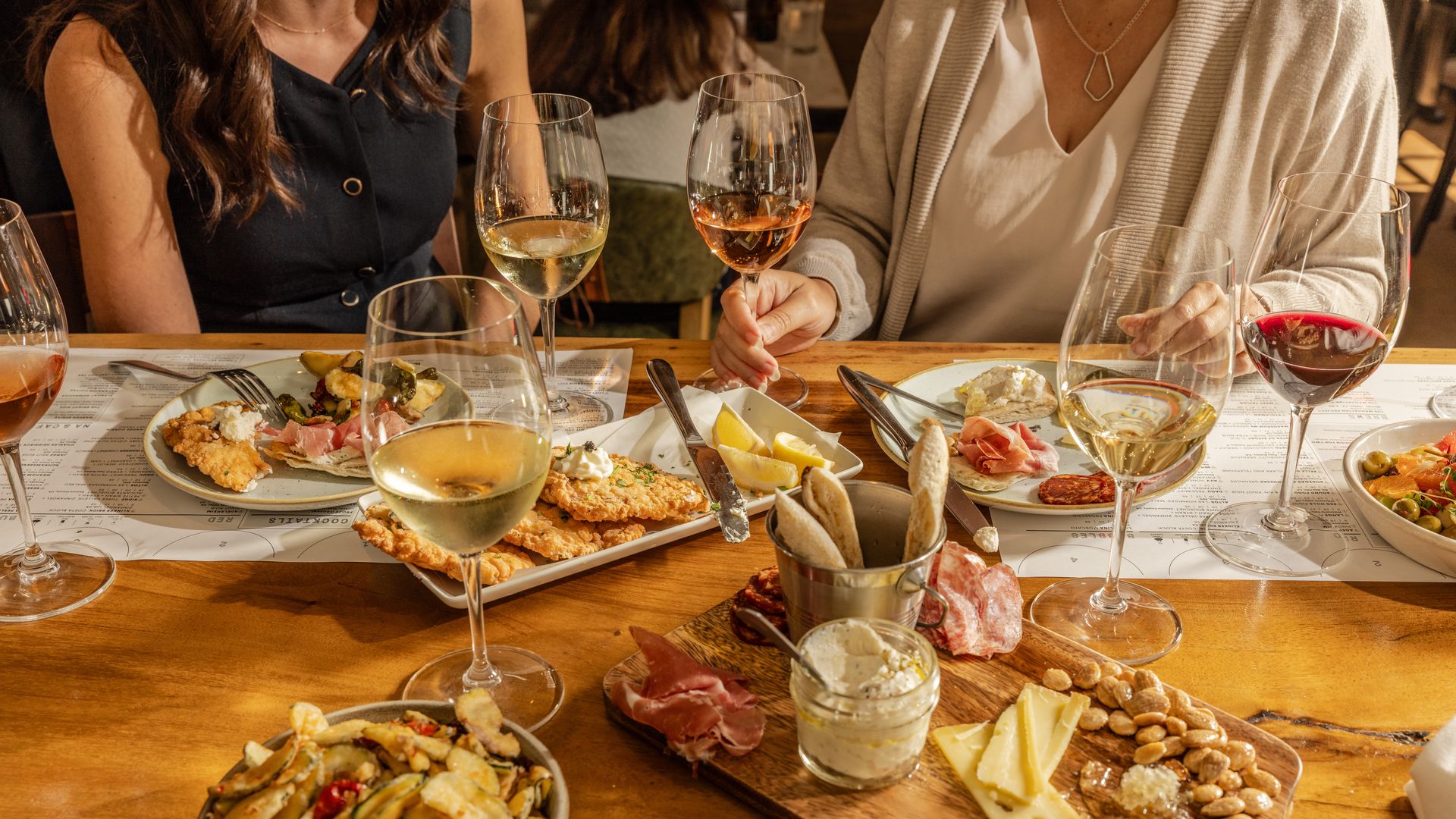 People enjoying a meal with various appetizers like cheese, nuts, cured meats, and fried snacks on a wooden table, accompanied by glasses of white, red, and rosé wine.