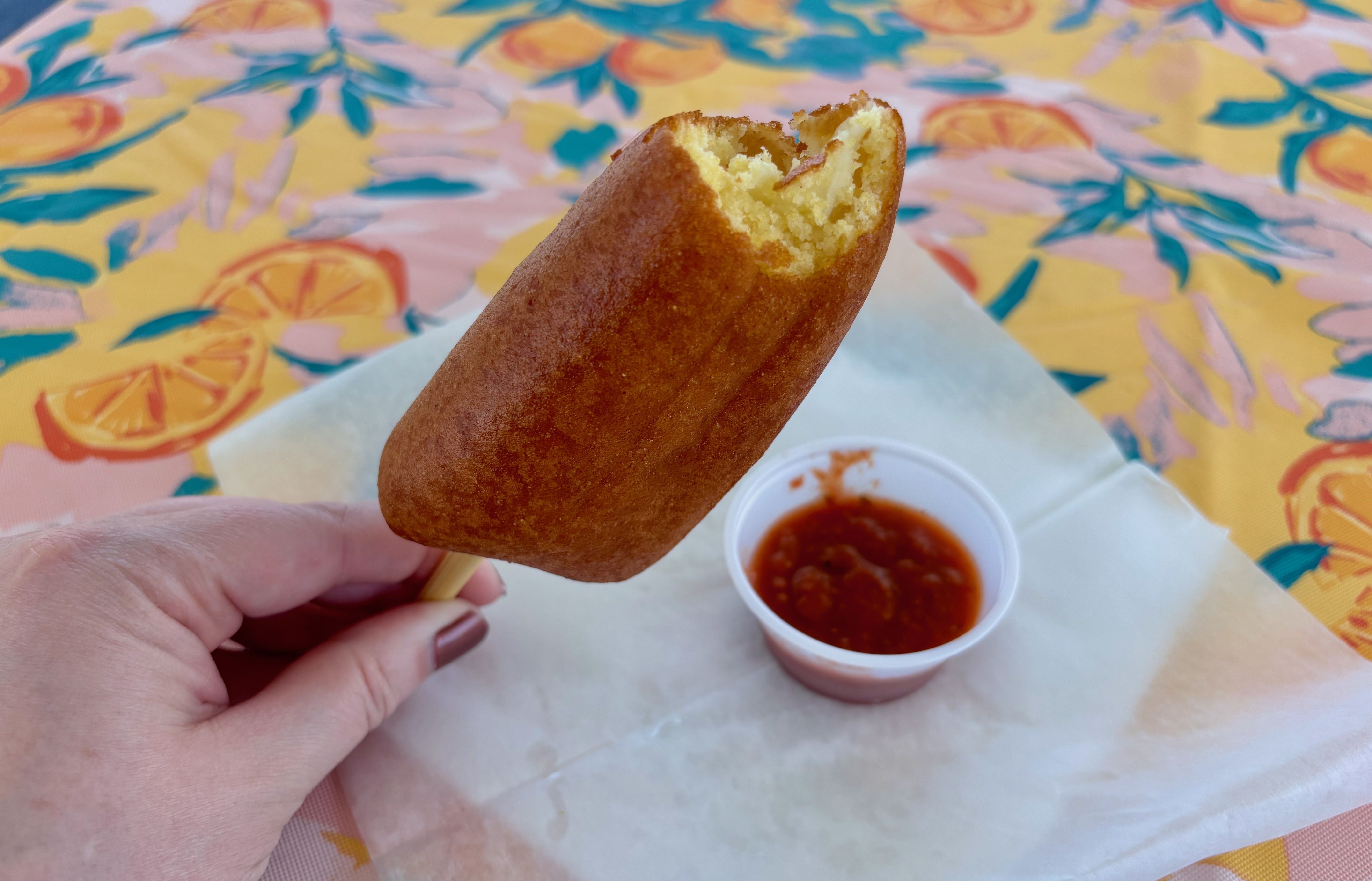 Hand holding a bitten fried cheese on a stick with corndog breading over wax paper, with a small cup of red dipping sauce on a yellow tablecloth with orange and green leaf patterns.