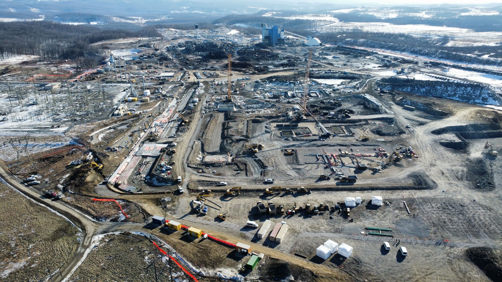 An aerial view of a large winter construction site with cranes, excavations, and trucks spread across muddy ground; distant snow-covered fields and wooded hills surround the site.