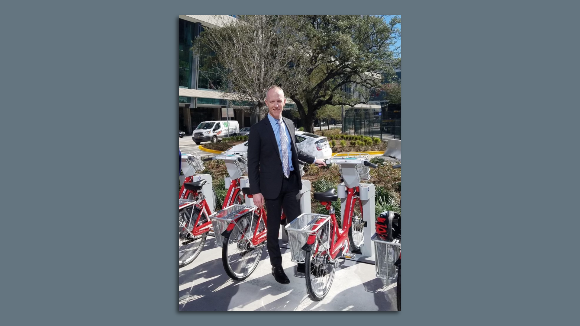 Pat Walsh, wearing a black suit with a blue shirt, poses for a photo next to a series of red BCycle bicycles 