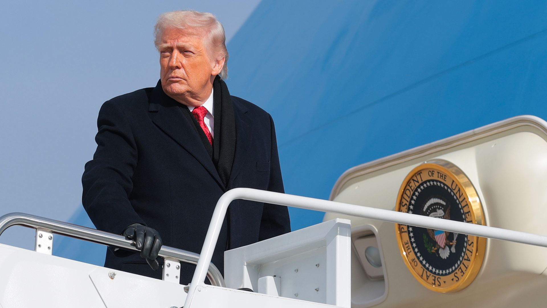 President Trump, in a suit with red tie and overcoat, looks over his shoulder as he boards Air Force One. 