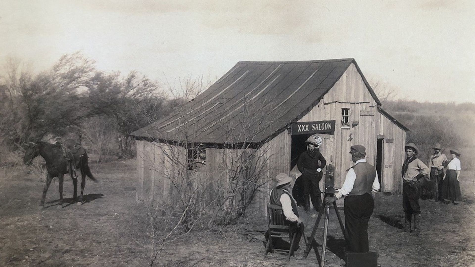 Black-and-white frontier scene: a wooden saloon shack labeled "XXX Saloon" with a metal roof. A man sits by a tripod camera while several men stand nearby; two horses are tied to a post on the left.