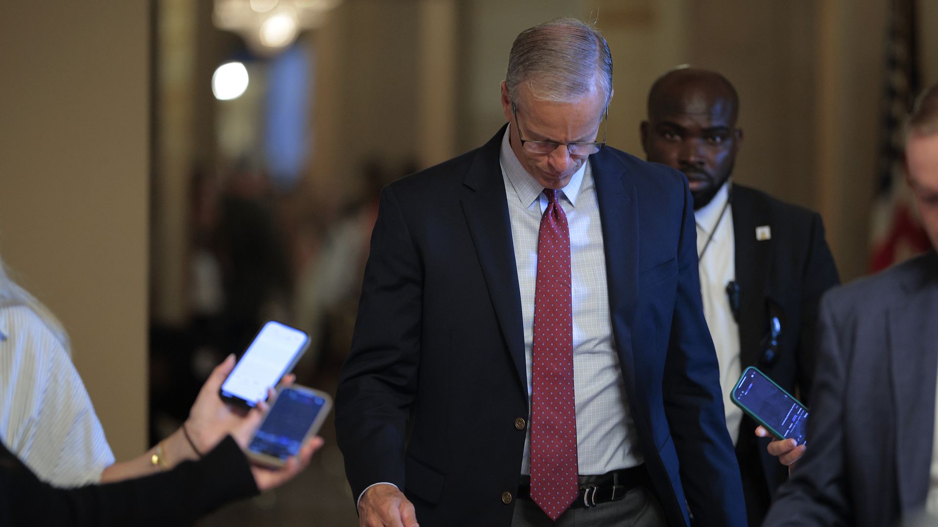 Man in a dark blue suit, light blue shirt, and red patterned tie looking down, surrounded by people holding smartphones in a hallway with blurred background.