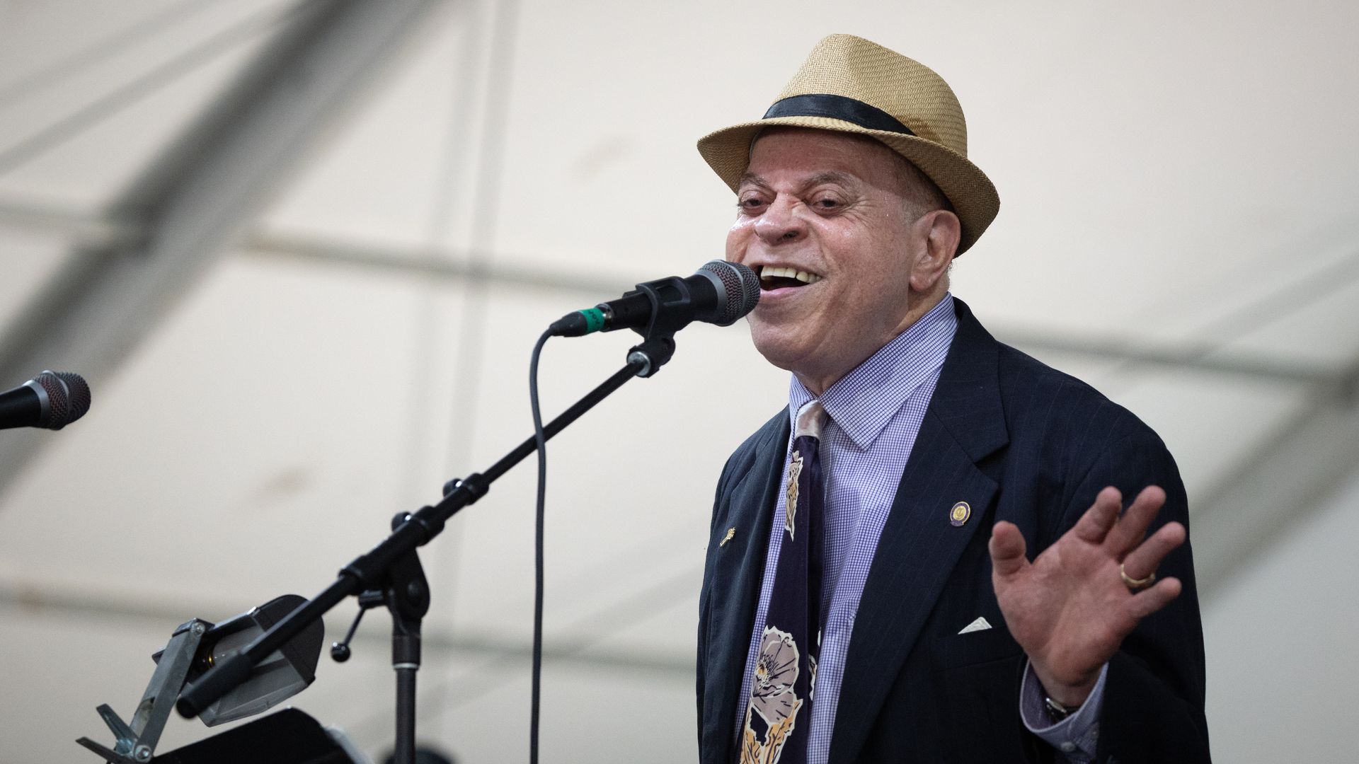 Deacon John sings into a microphone and holds one hand up.
