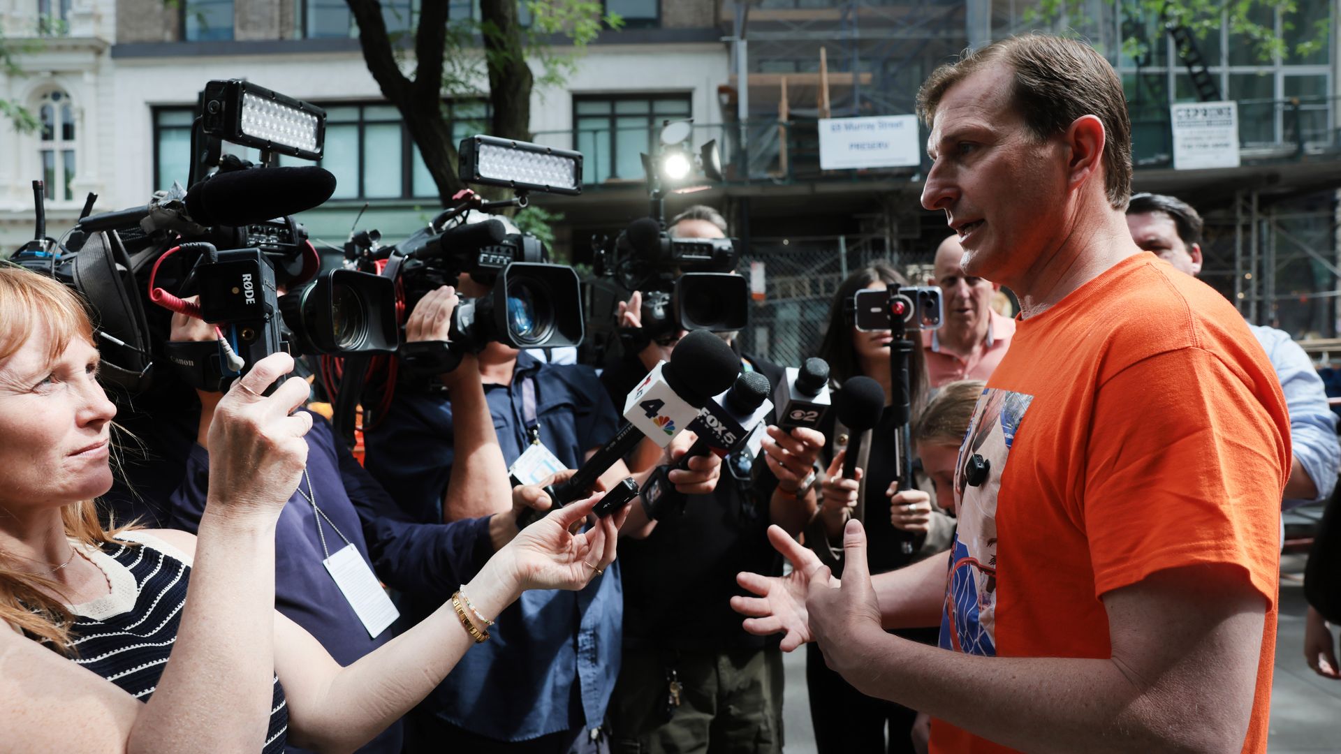 Man in an orange shirt speaks to a crowd of reporters with microphones and cameras on a city street, surrounded by journalists; trees and buildings in the background.