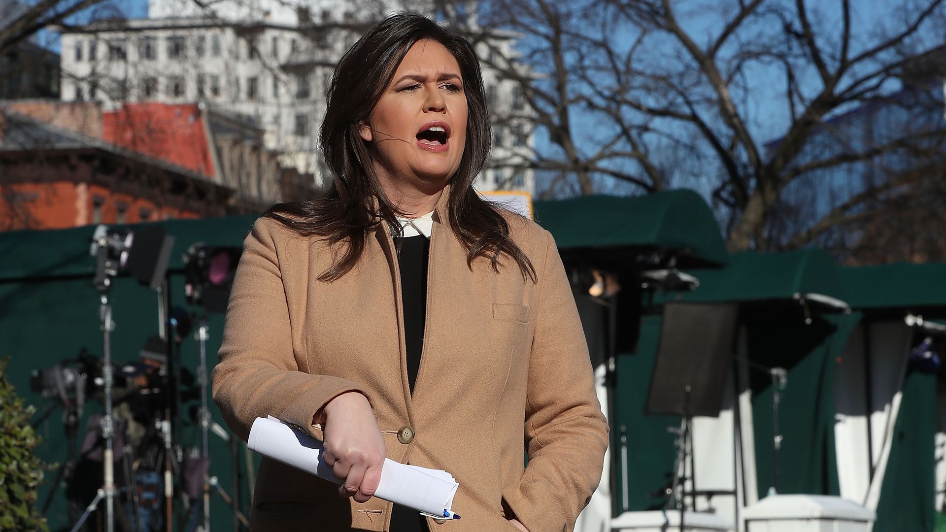 Sarah Sanders talking to reporters with a beige coat on.