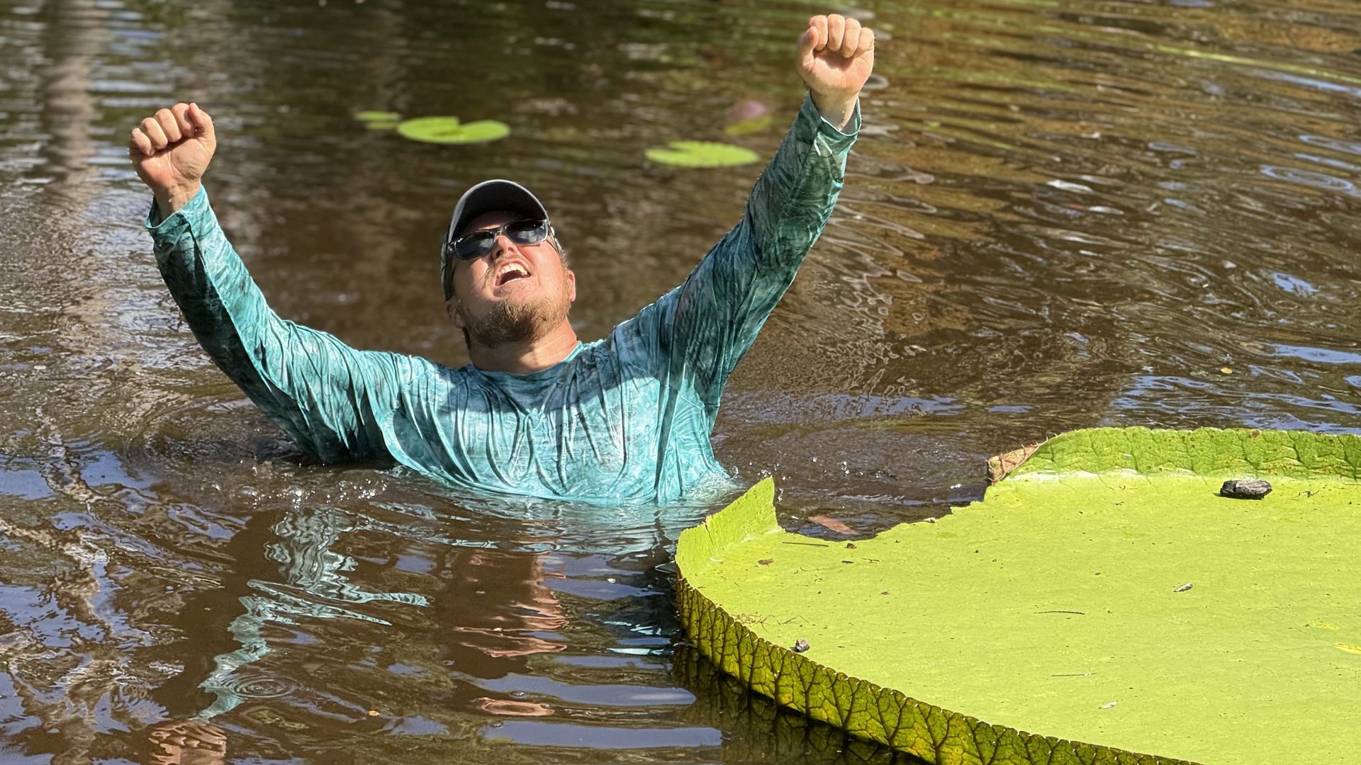 Man wearing a teal long-sleeve shirt, sunglasses, and a cap raises his fists while standing waist-deep in brown water near large green lily pads under sunlight.