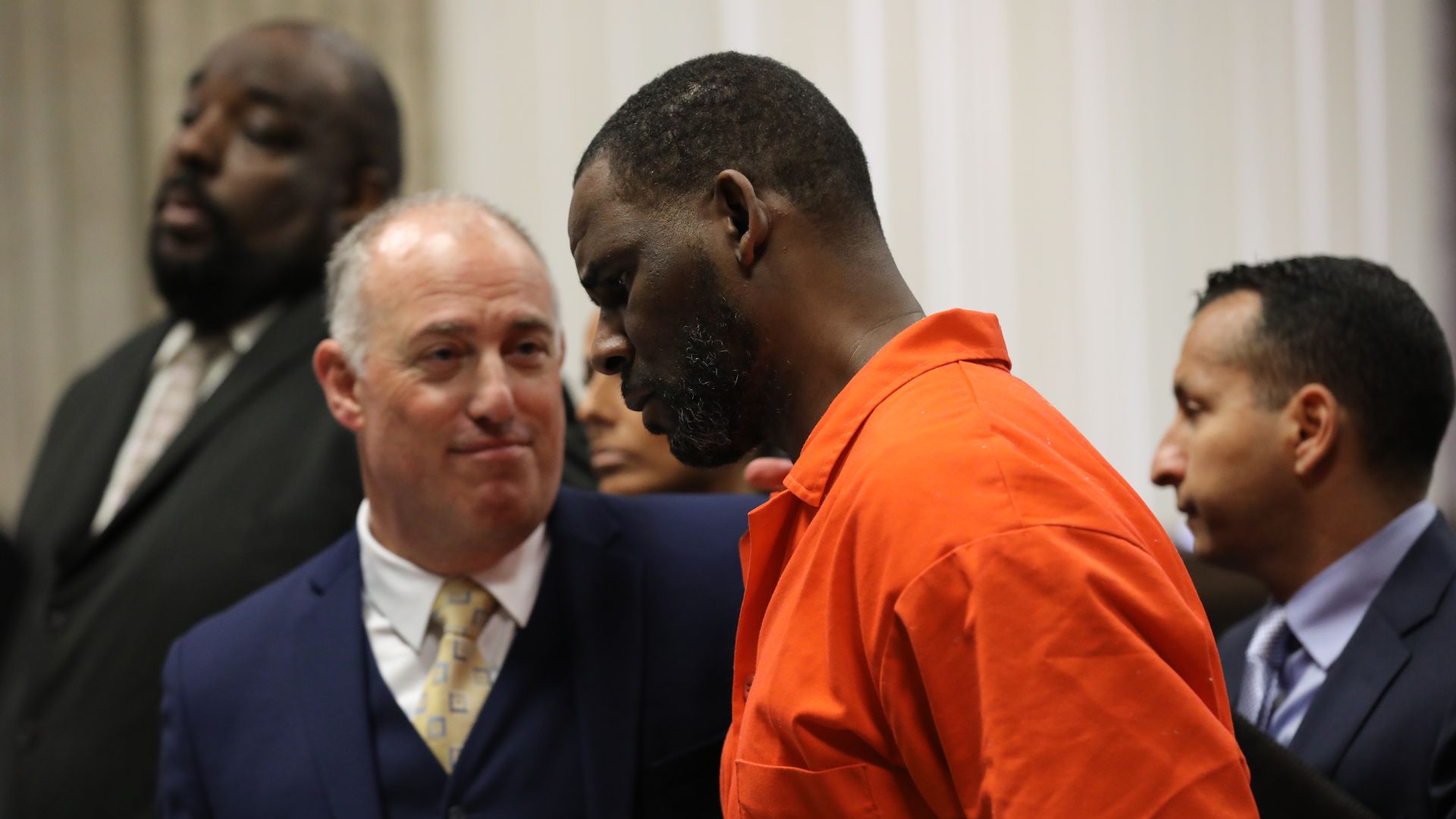 In this image, Kelly stands in an orange prison jumpsuit next to his lawyer, who wears a suit and tie.