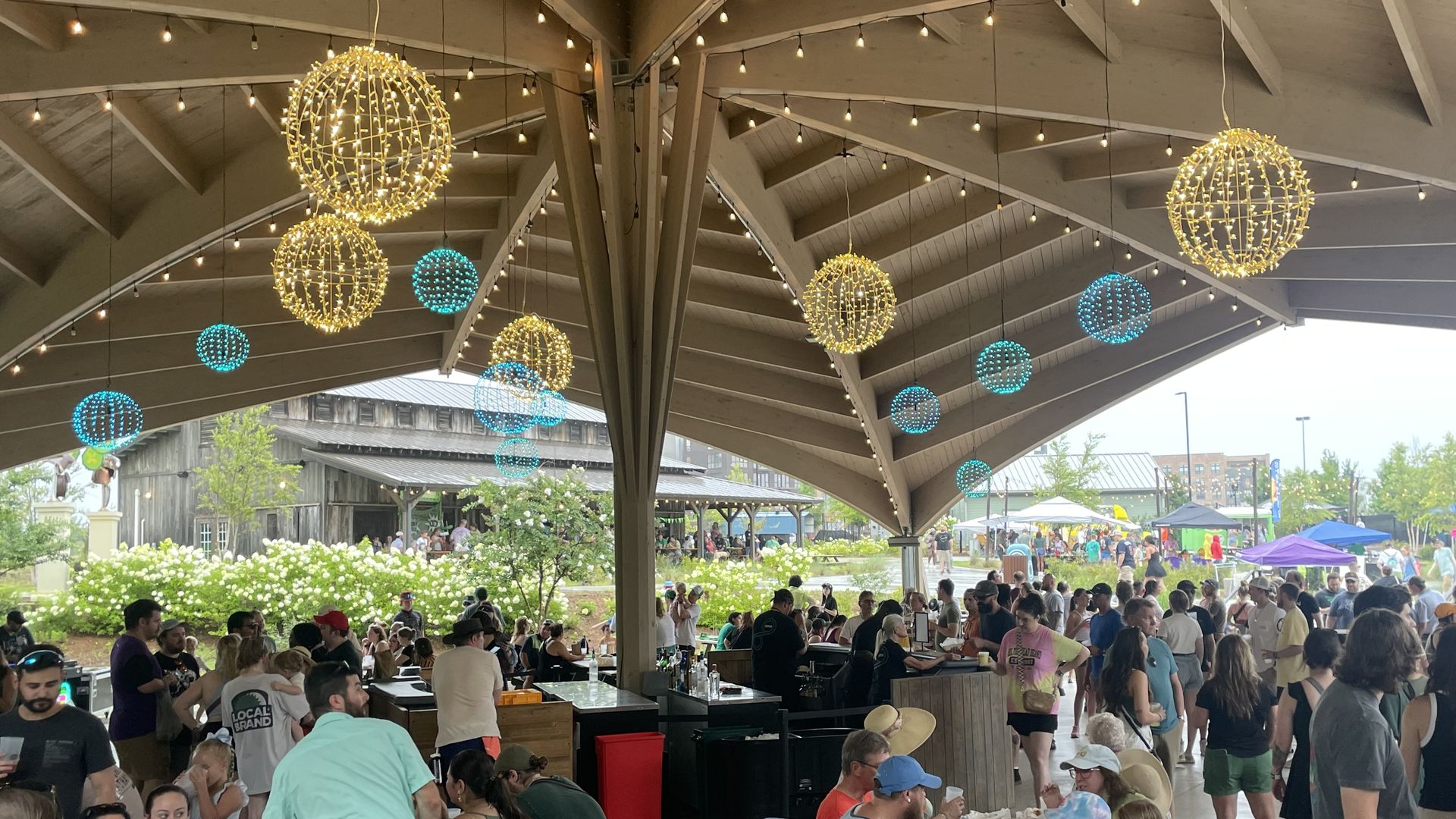 Crowded outdoor festival under a sheltered pavilion with hanging string lights and glowing gold and teal spherical lanterns; people sit at long tables eating as vendors line the edges outside.