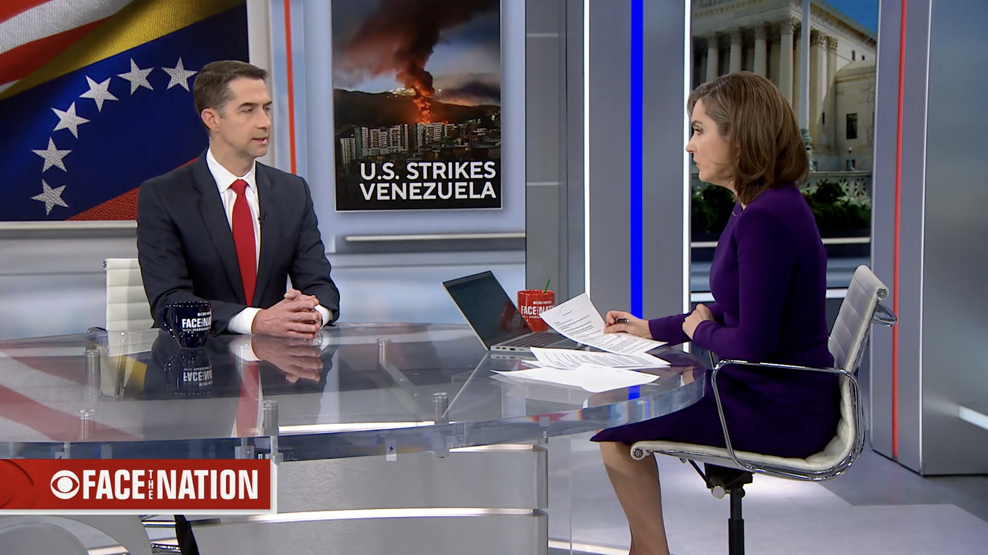 Senator Tom Cotton speaks with host Margaret Brennan on CBS's Face the Nation. A screen behind them shows an image of a burning city with the headline "U.S. Strikes Venezuela" next to the Venezuelan flag. Both are seated at a glass table with notes and coffee mugs.