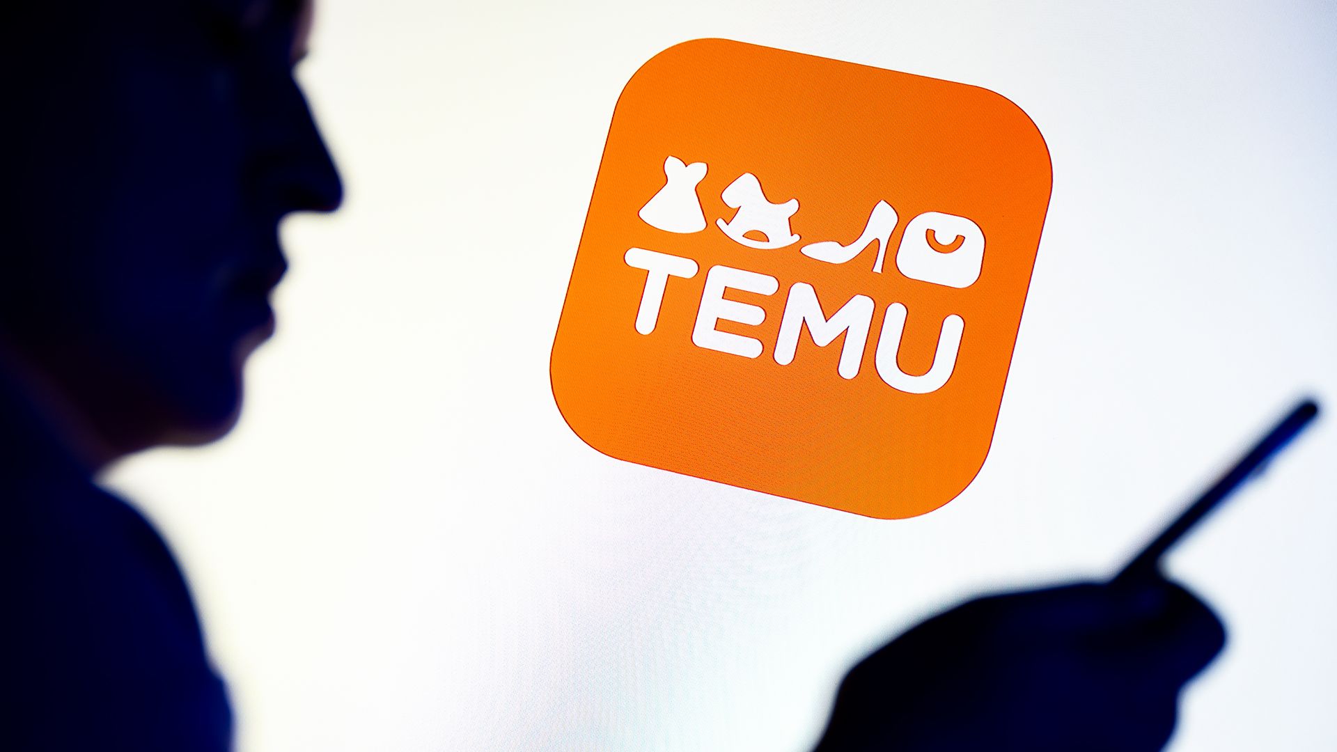 Silhouette of a person holding a phone next to a bright screen displaying the orange Temu app logo featuring icons of a dress, rocking horse, high heel, and shopping bag.