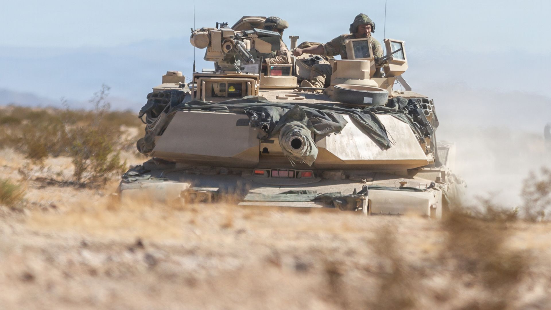 Desert-colored military tank with two soldiers visible inside, moving through dry, sandy terrain under a clear sky.