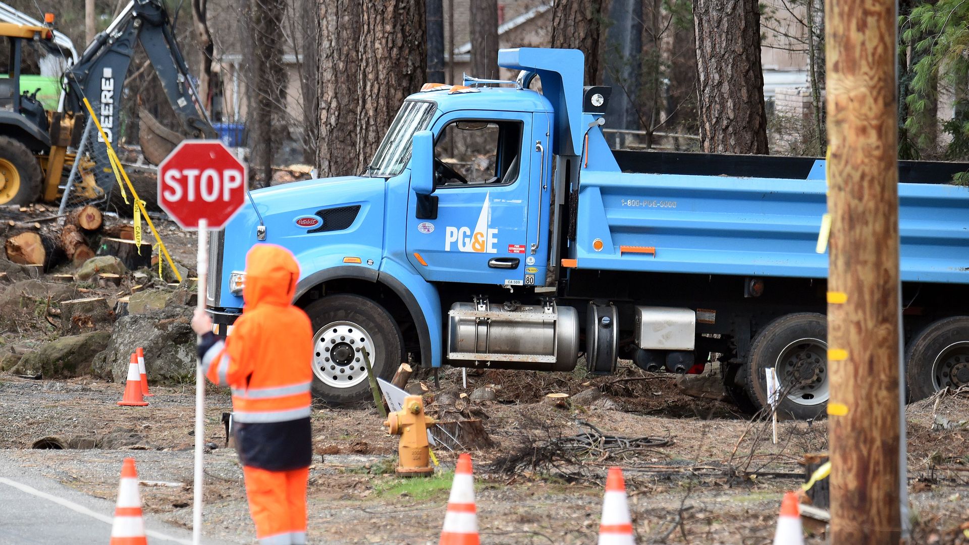 A construction worker holds a stop sign in front of a blue PG&E service vehicle in a burned area.