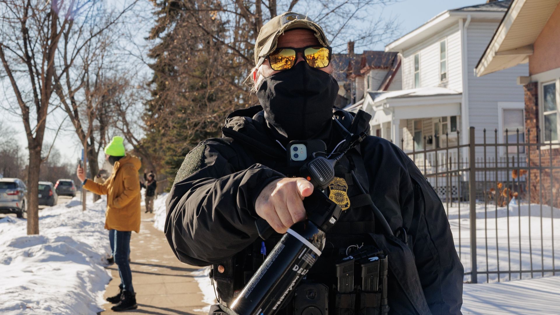 A federal agent in black gear, mask, and reflective sunglasses pointing while holding a black canister, standing on a snowy sidewalk with a person in a yellow jacket and neon green hat using a phone behind them.
