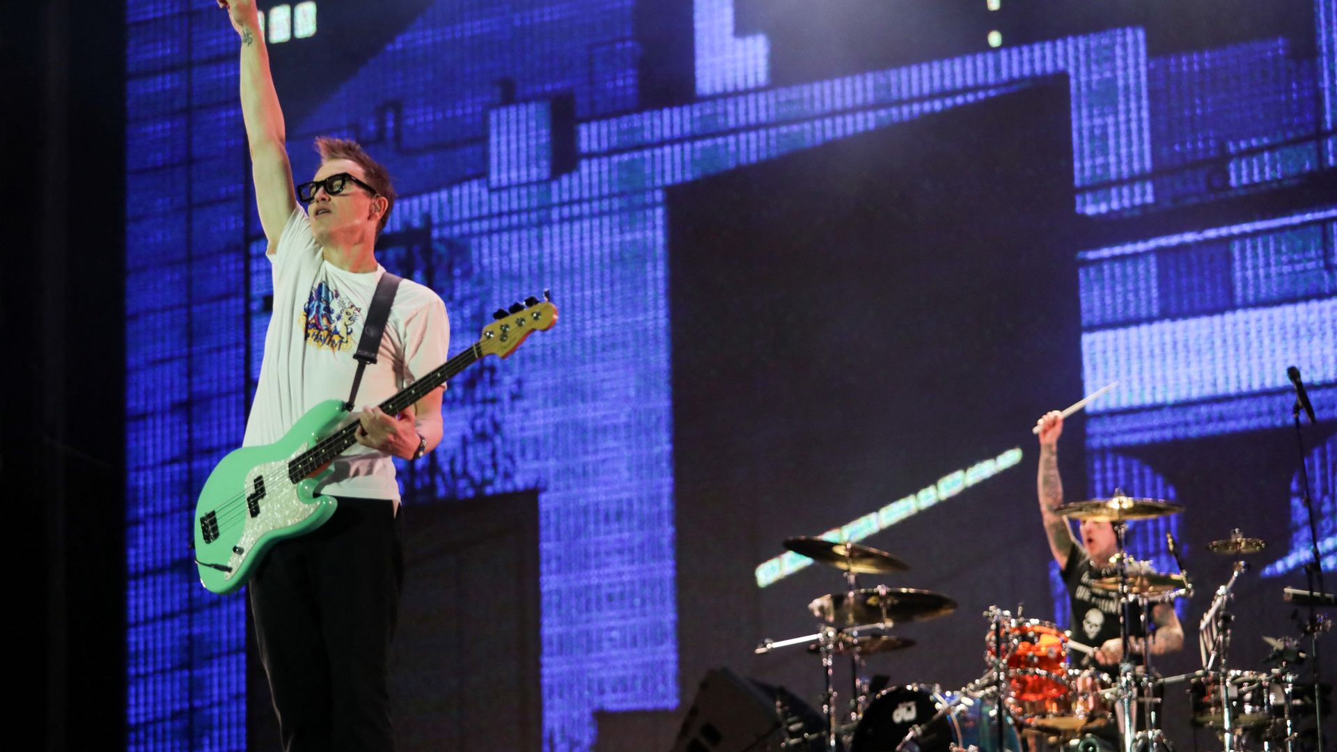 A rock guitarist and drummer perform on a stage in front of a large digital screen. The guitarist is wearing sunglasses and hold up their arm in the air to interact with the crowd.