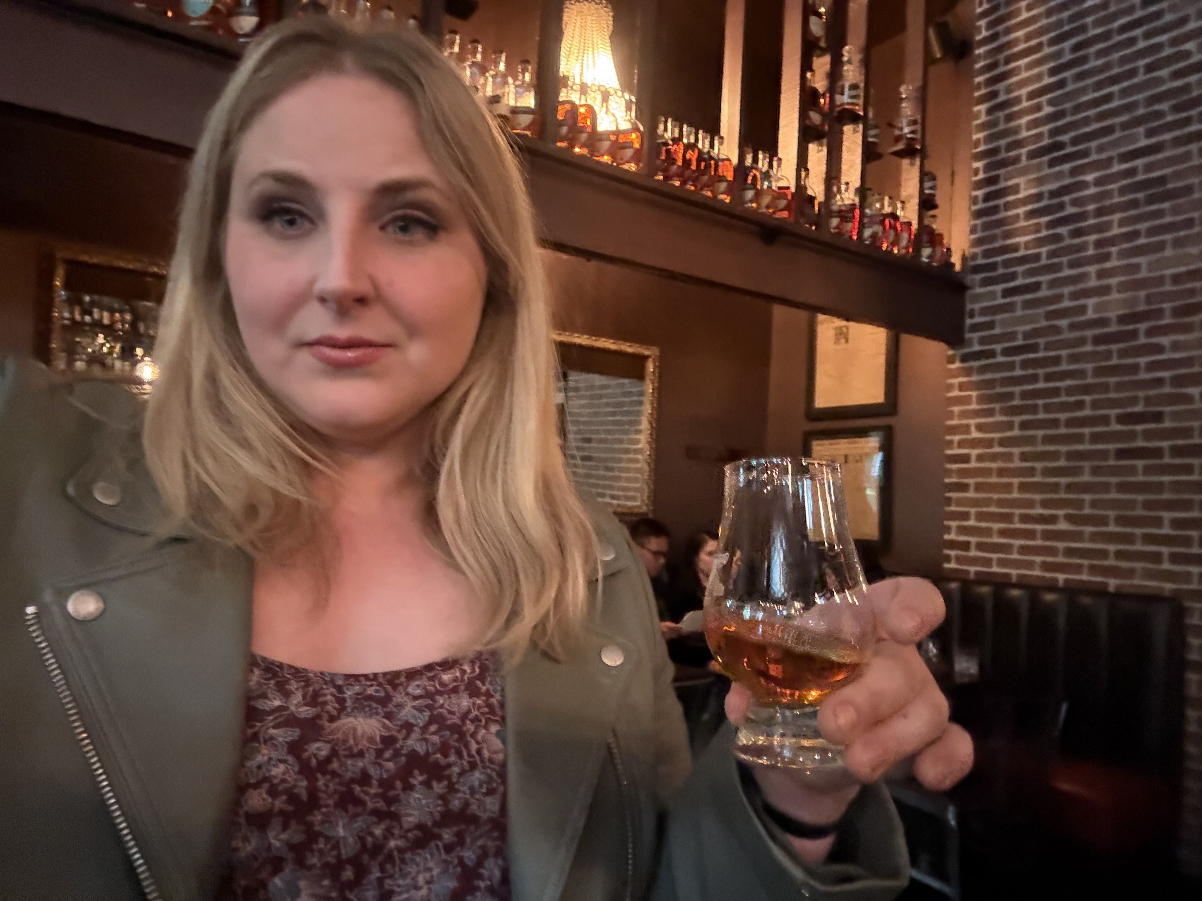 Blonde woman in a green jacket holds an amber whiskey glass in a dim bar. Warm light, brick wall, and bottles on shelves glow behind her.
