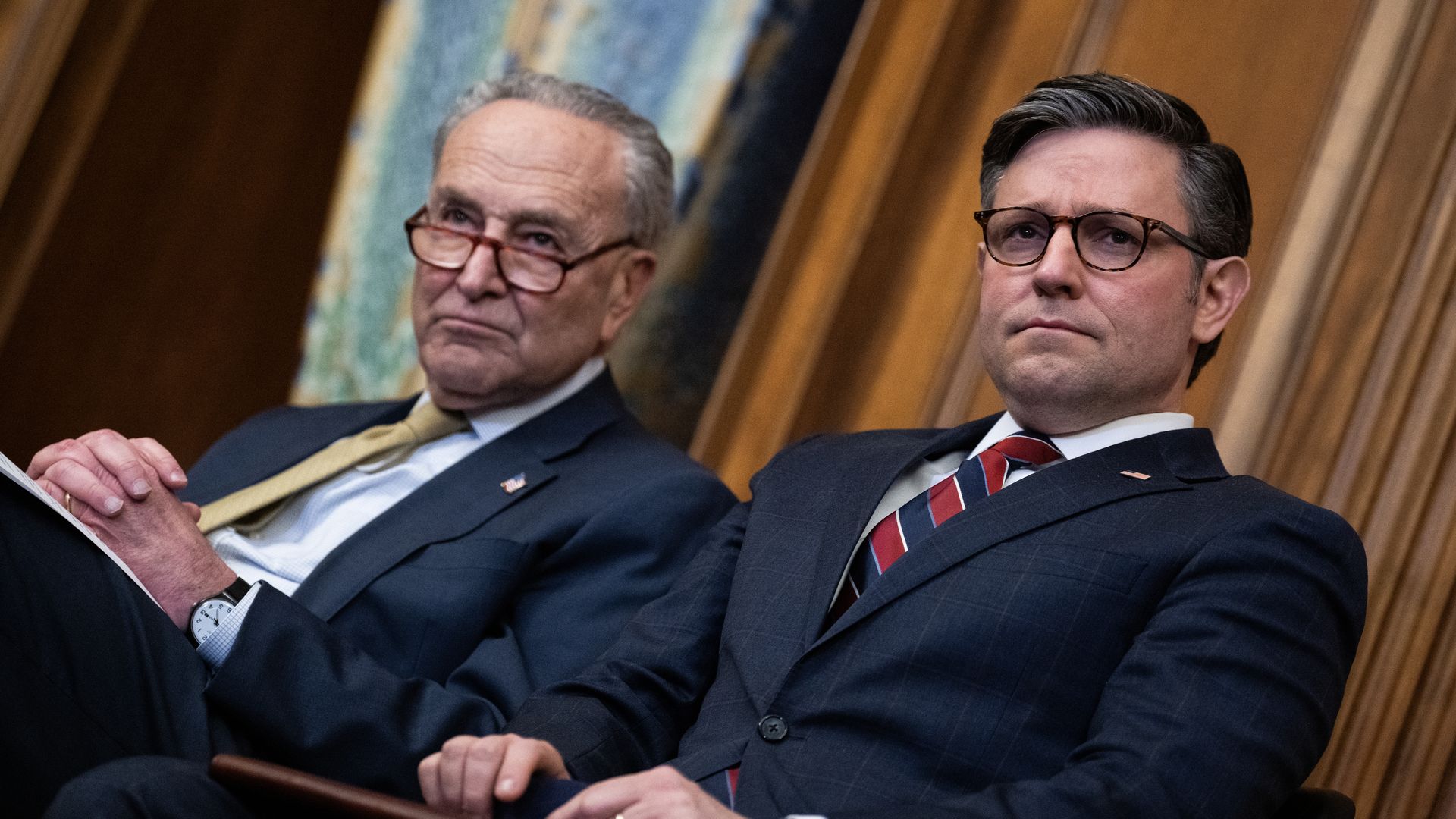 Senate Majority Leader Schumer, wearing a dark blue suit, light blue shirt and gold tie, sits next to House Speaker Mike Johnson, wearing a dark blue suit, light blue shirt and red and blue striped tie, both wearing glasses and seated in front of a wood-paneled wall.