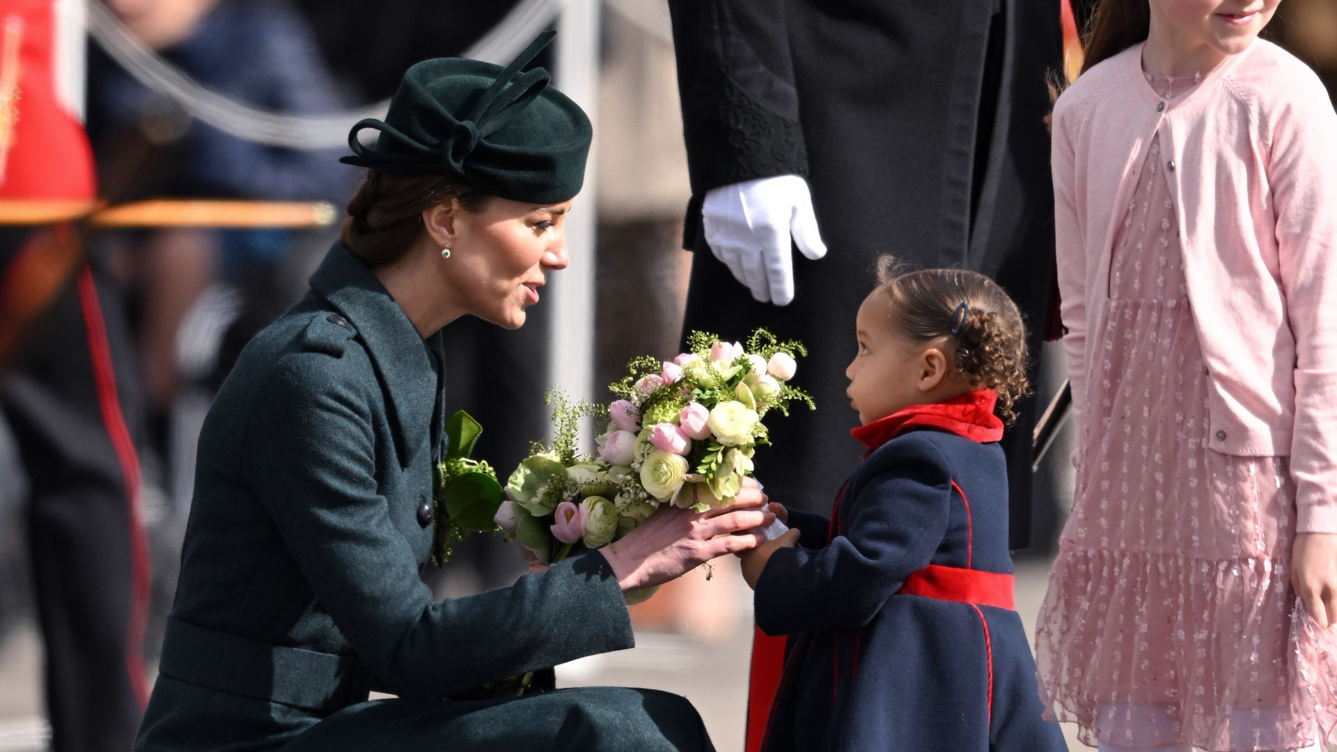 Kate Middleton, the Duchess of Cambridge, is attired in a black hat and coat as she receives white flowers from a young girl in a black and red coat.