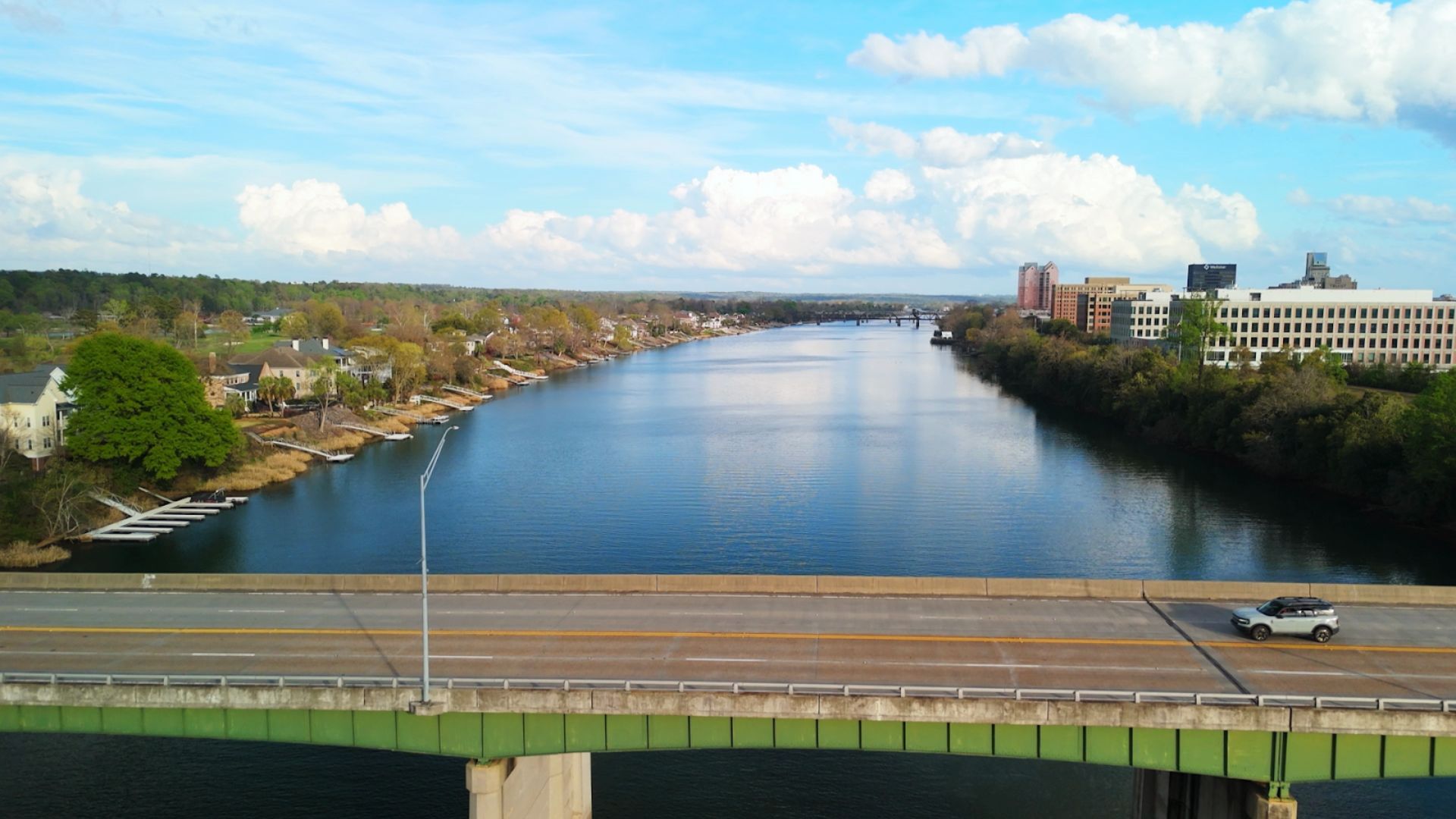 Aerial view of a wide river between a tree-lined suburban shore with docks on the left and a modern office complex on the right; a green bridge and highway cross the foreground with a white SUV.