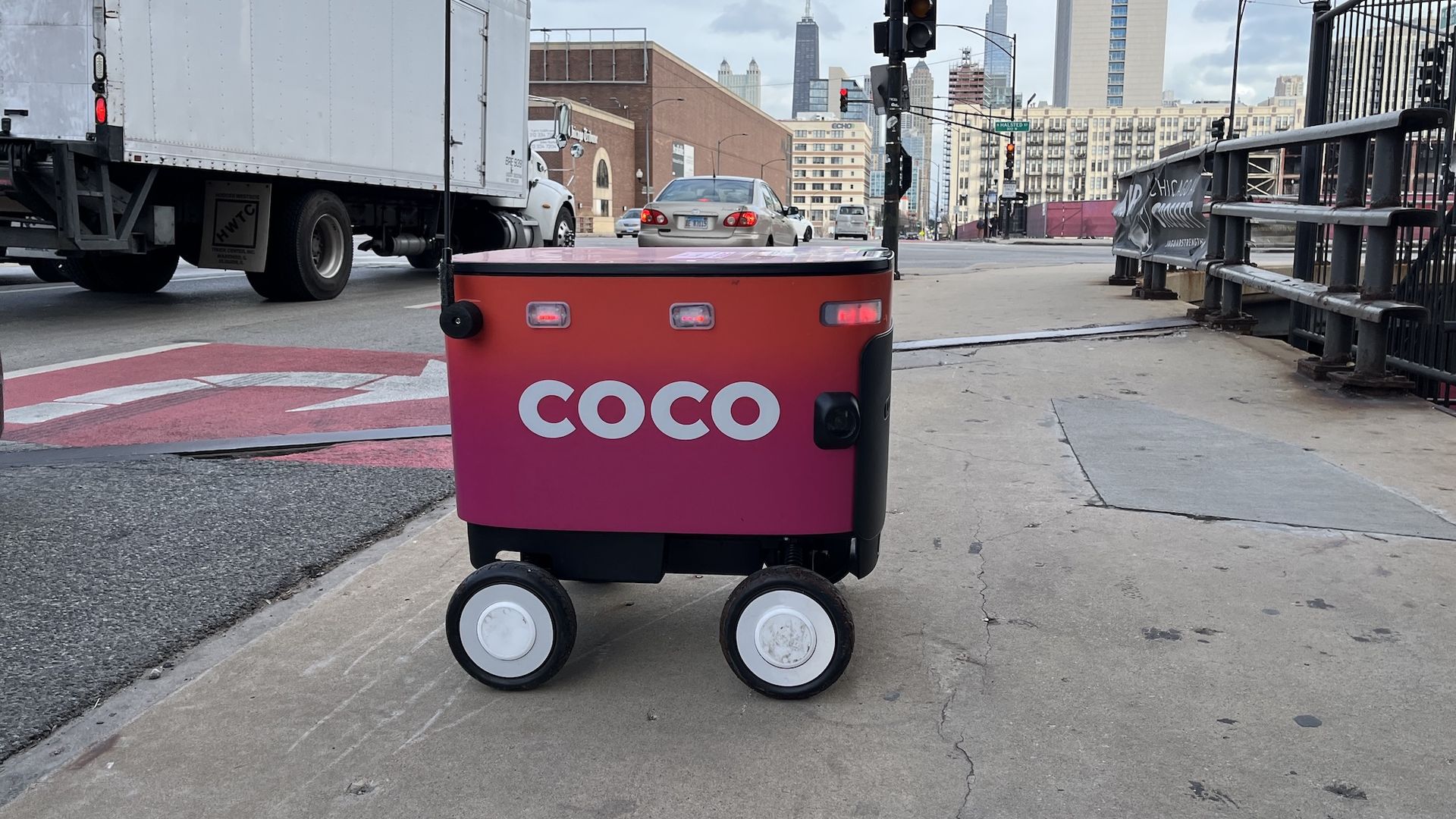 Pink-to-orange gradient COCO delivery robot on four white wheels sits on a city sidewalk near a crosswalk, with a red traffic light, trucks, and tall city buildings in the background.