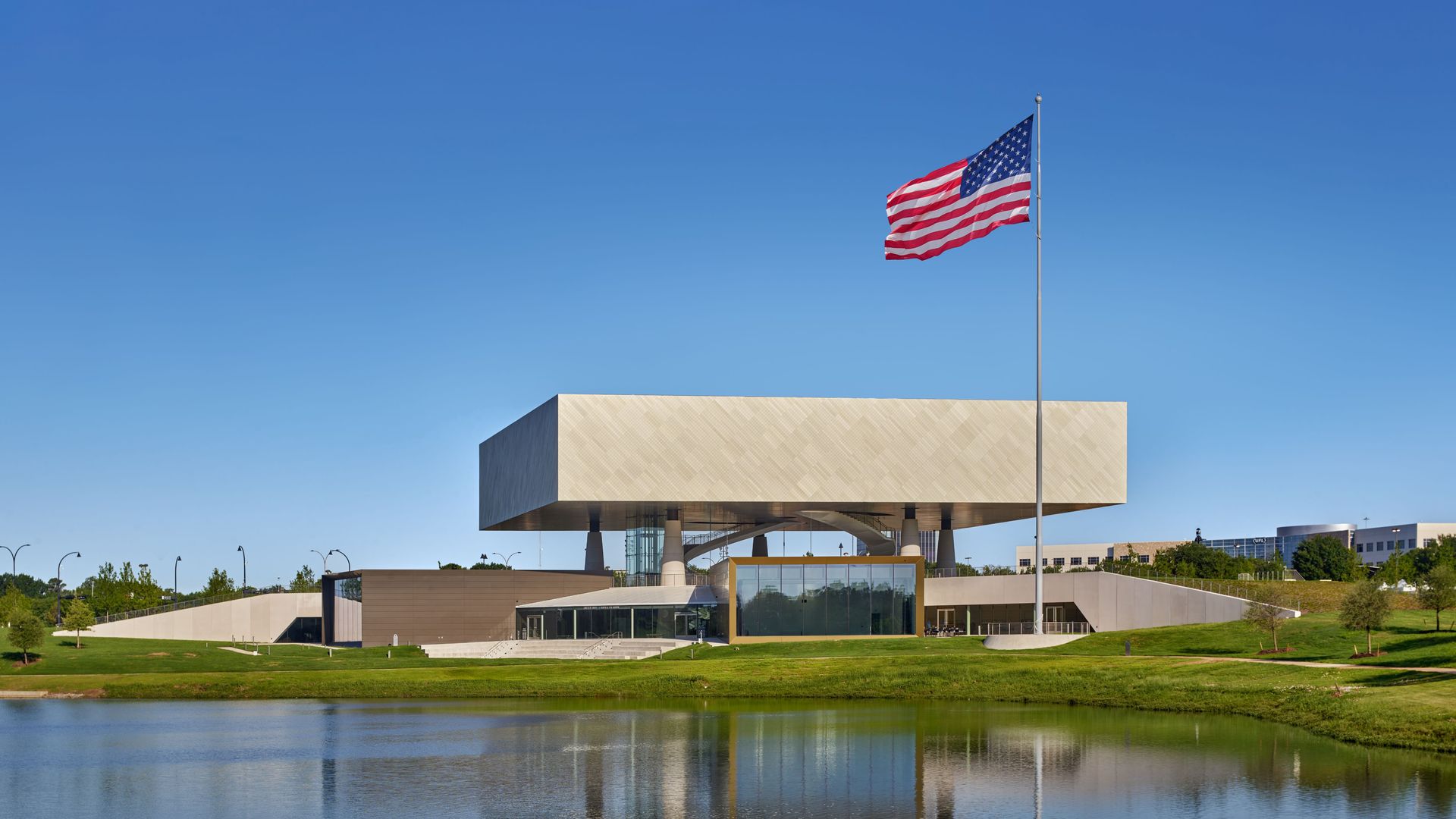 Modern rectangular building with large American flag on flagpole, grassy area, and pond reflecting the scene under a clear blue sky.