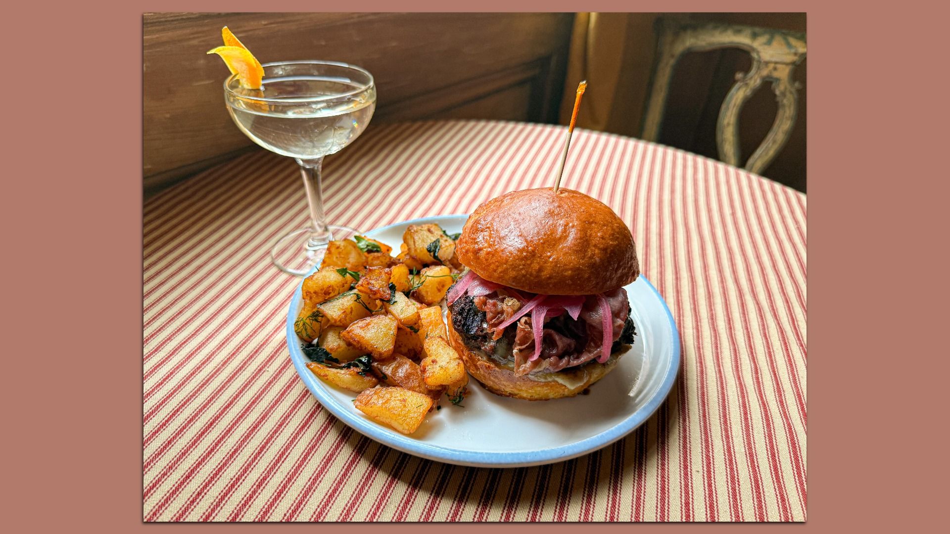 Plate with a burger topped with pink pickled onions and a side of golden roasted potatoes on a red and white striped tablecloth, next to a cocktail glass with an orange peel garnish.