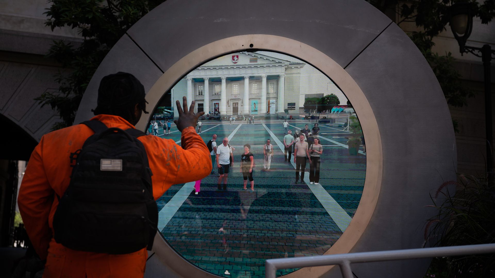 Back view of a person in an orange jacket with a backpack touching a circular glass screen that shows a crowded plaza and a white neoclassical building with columns, framed by a metal sculpture.