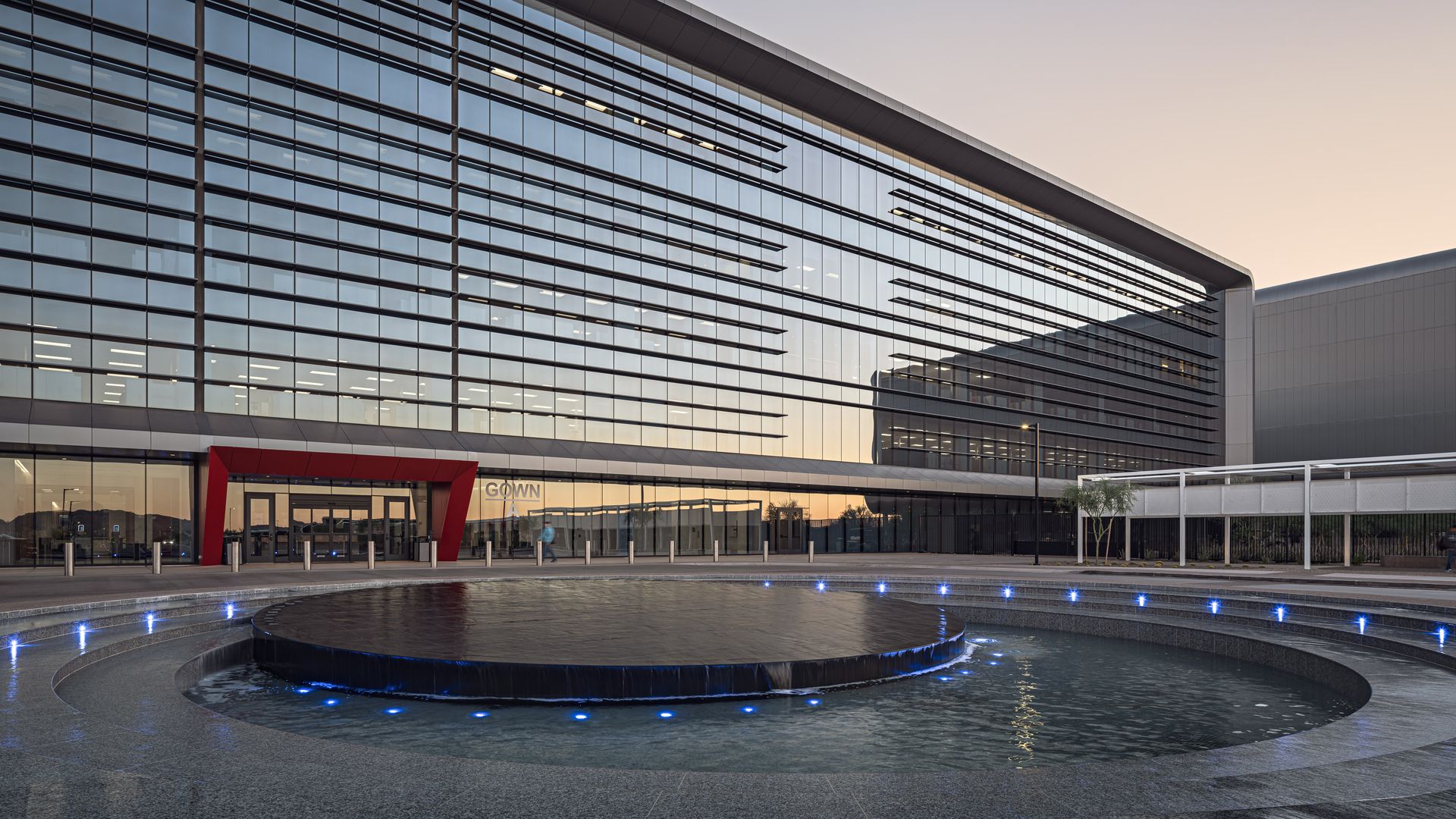Modern glass building with horizontal lines and a red-framed entrance, reflected sky at dusk, and circular water fountain with blue lights in front.