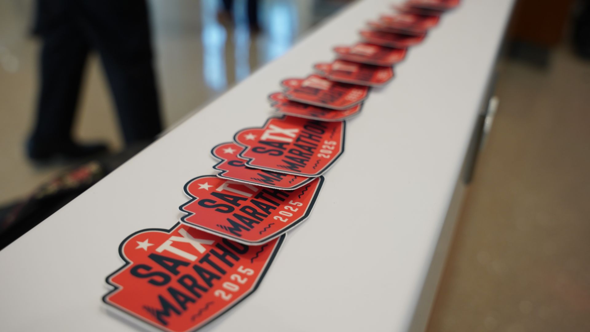 Red and black stickers arranged in a line on a white surface, each reading "SATX MARATHON 2025," with blurred background showing people standing indoors.
