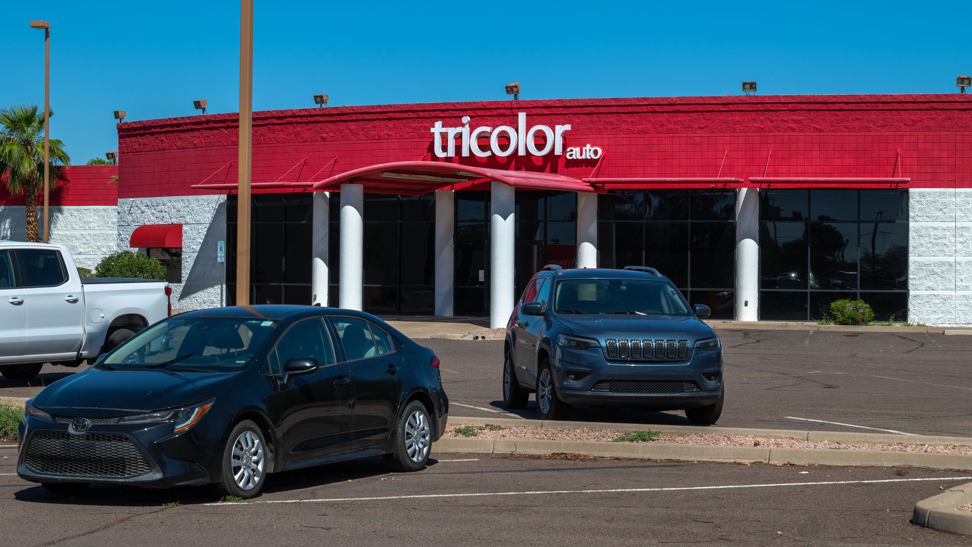 Black sedan and dark SUV parked in front of a white and red building with "tricolor auto" sign under a clear blue sky.