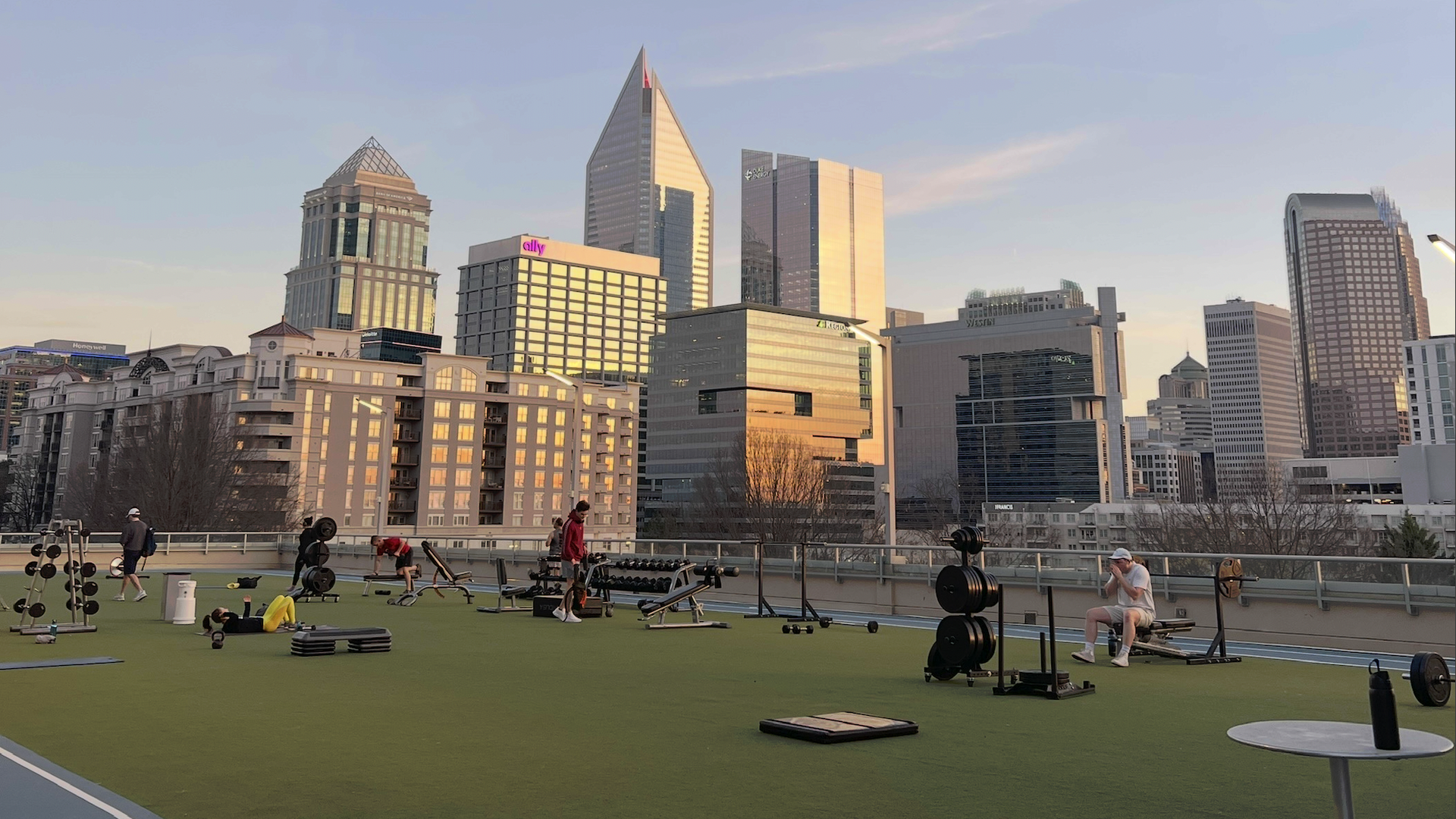 Rooftop gym on a city skyline: people lifting weights on green turf with benches and dumbbells, overlooking tall modern buildings bathed in sunset light.
