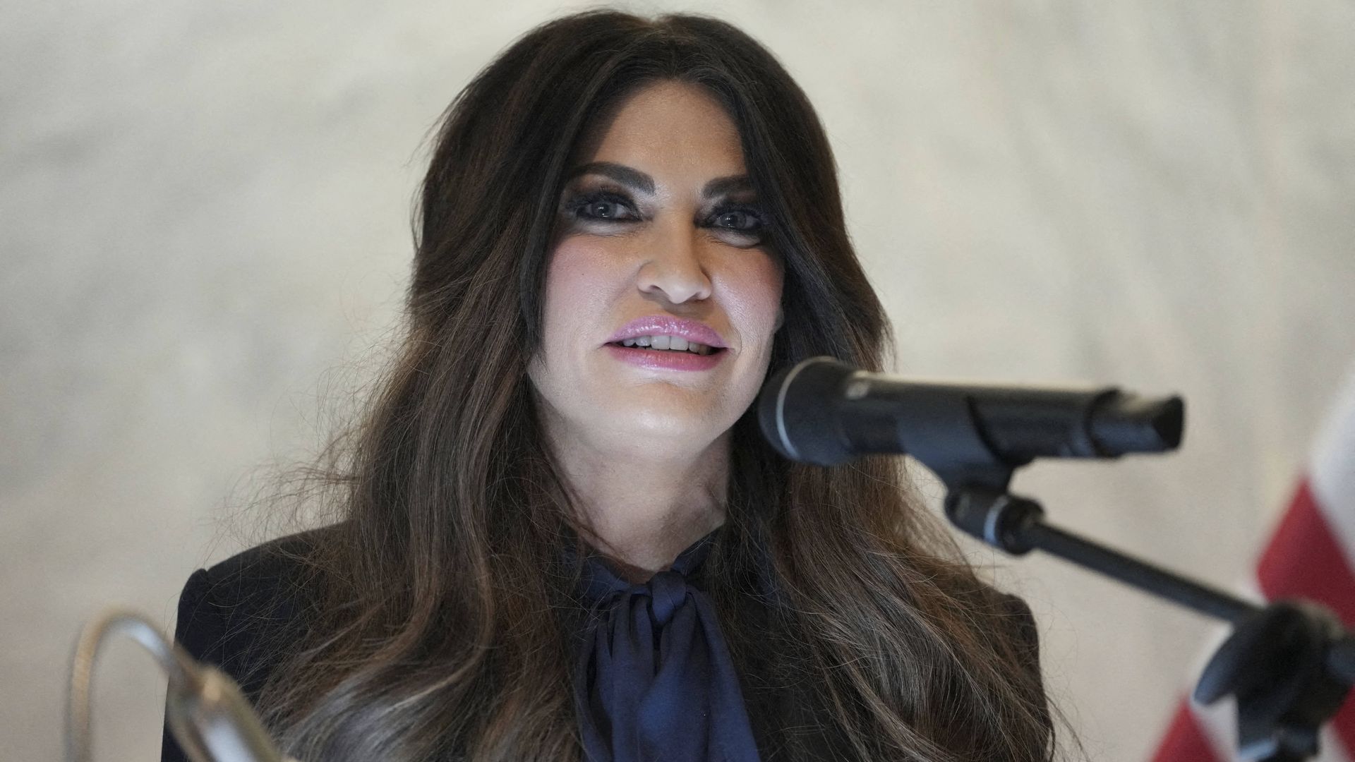 U.S. Ambassador to Greece Kimberly Guilfoyle, pale with dark hair and large, pink-painted lips and wearing a navy blouse, stares ahead with her green eyes beside a black mic.