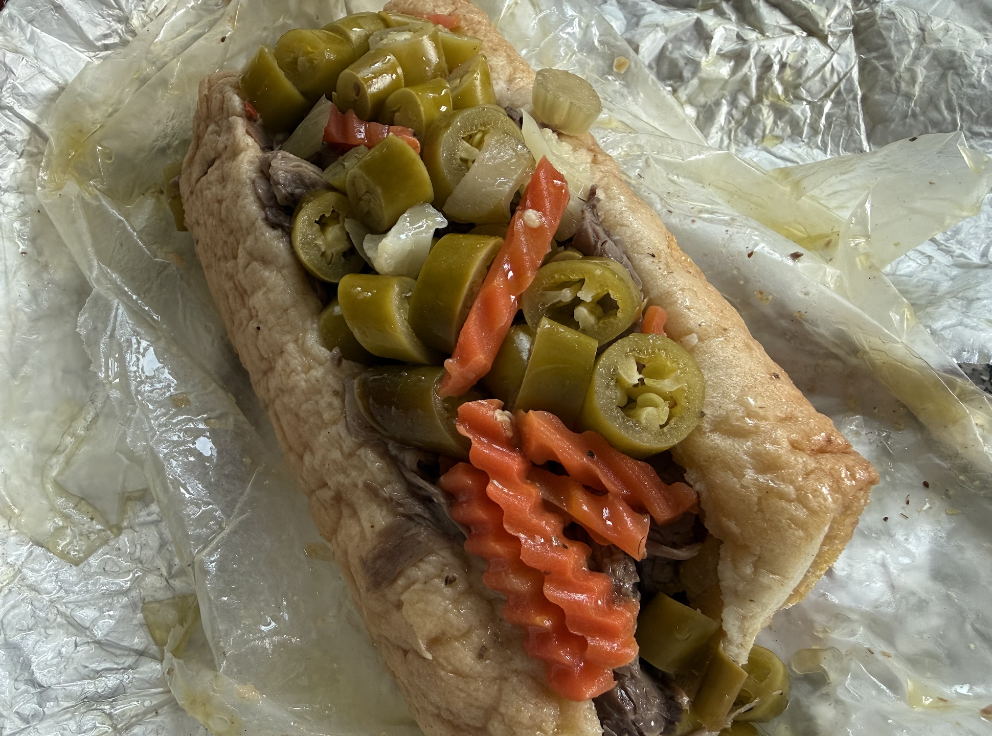 Close-up of a hot dog in a bun topped with chopped pickles, onion chunks, and orange crinkle-cut carrot strips, all on a foil-lined surface.
