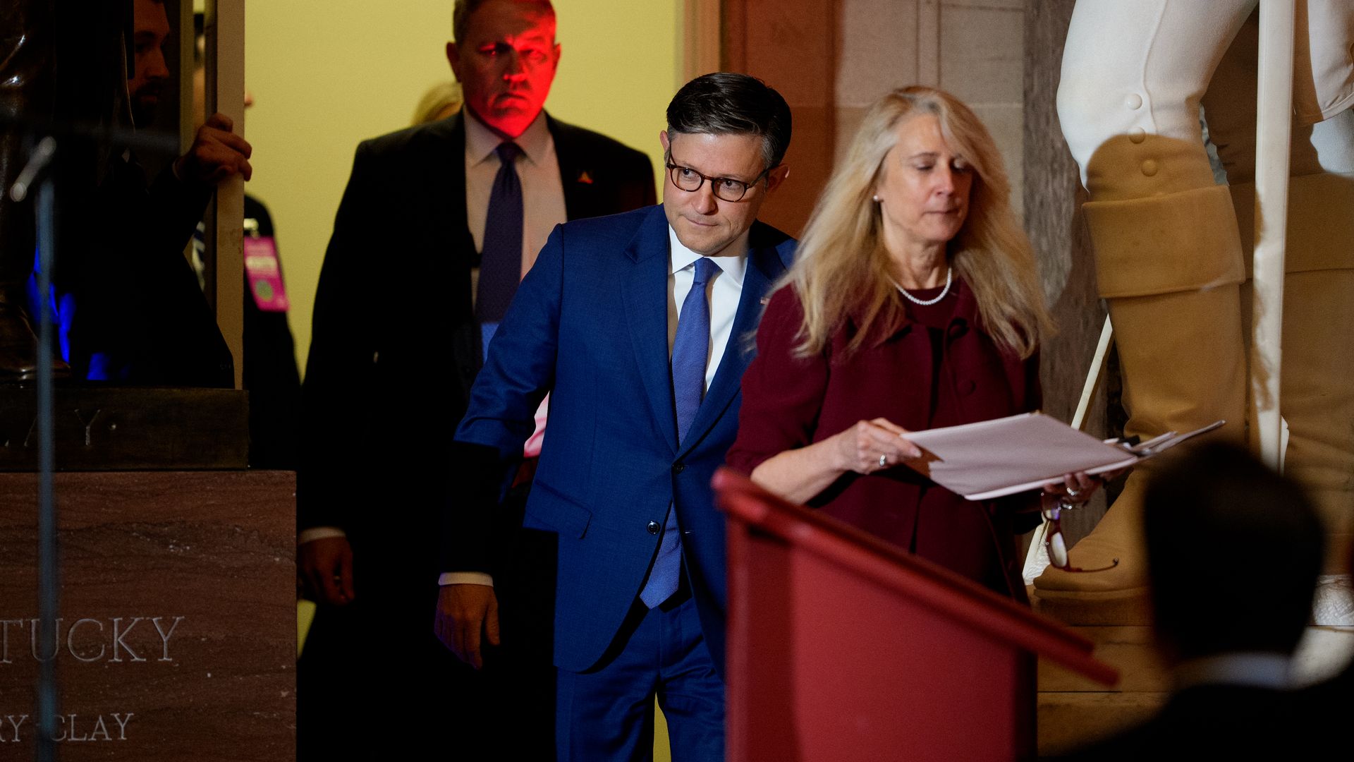 Two formally dressed people walk through a lobby; a man in a blue suit and glasses looks down while a woman in burgundy reads papers. A large statue leg stands on the right against a yellow wall.