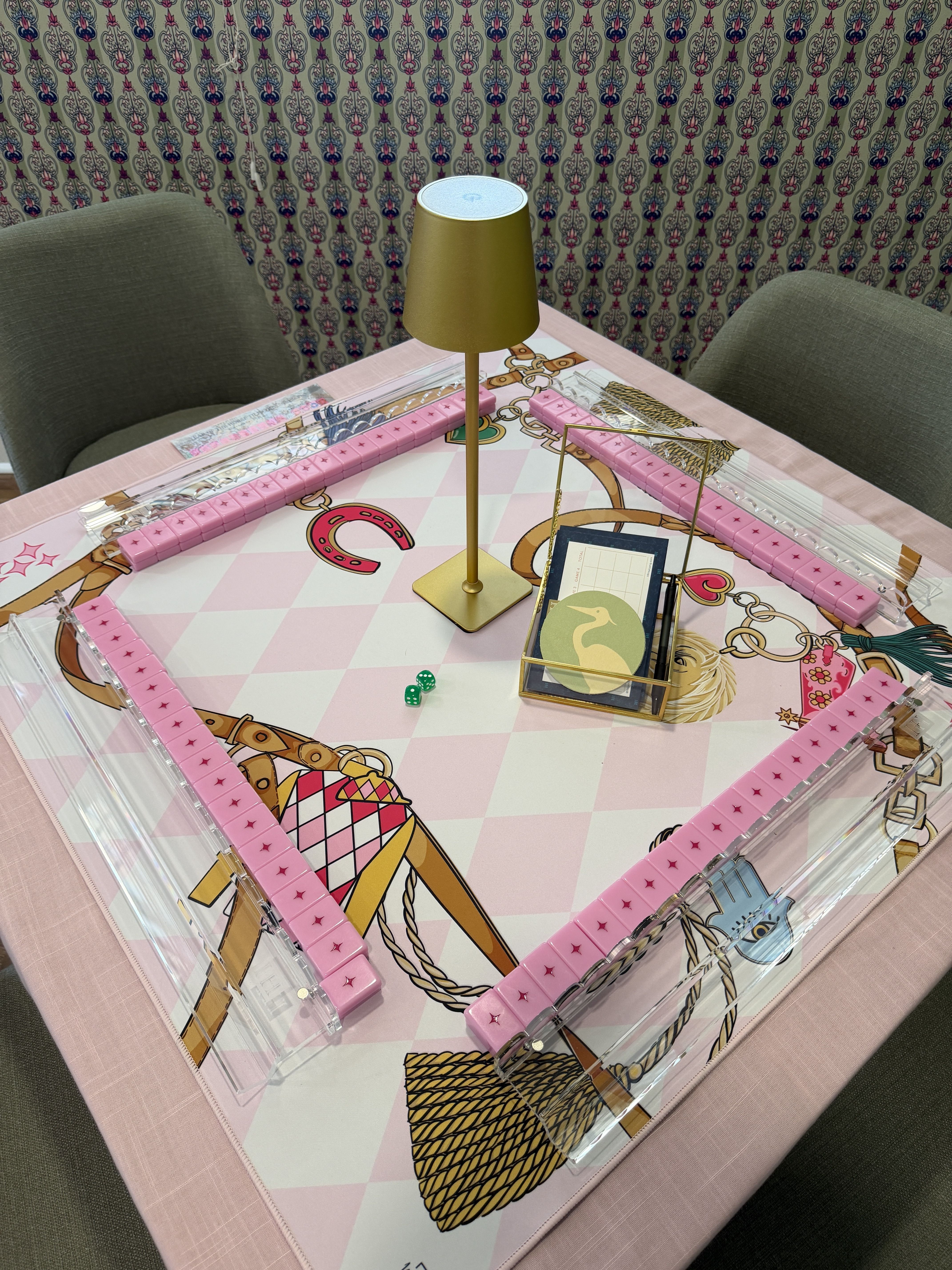 Pastel pink board game table with a tall gold lamp in the center, clear acrylic rails, and pink edge pieces. Two gray chairs sit beside a patterned wallpaper; scattered game pieces and dice.