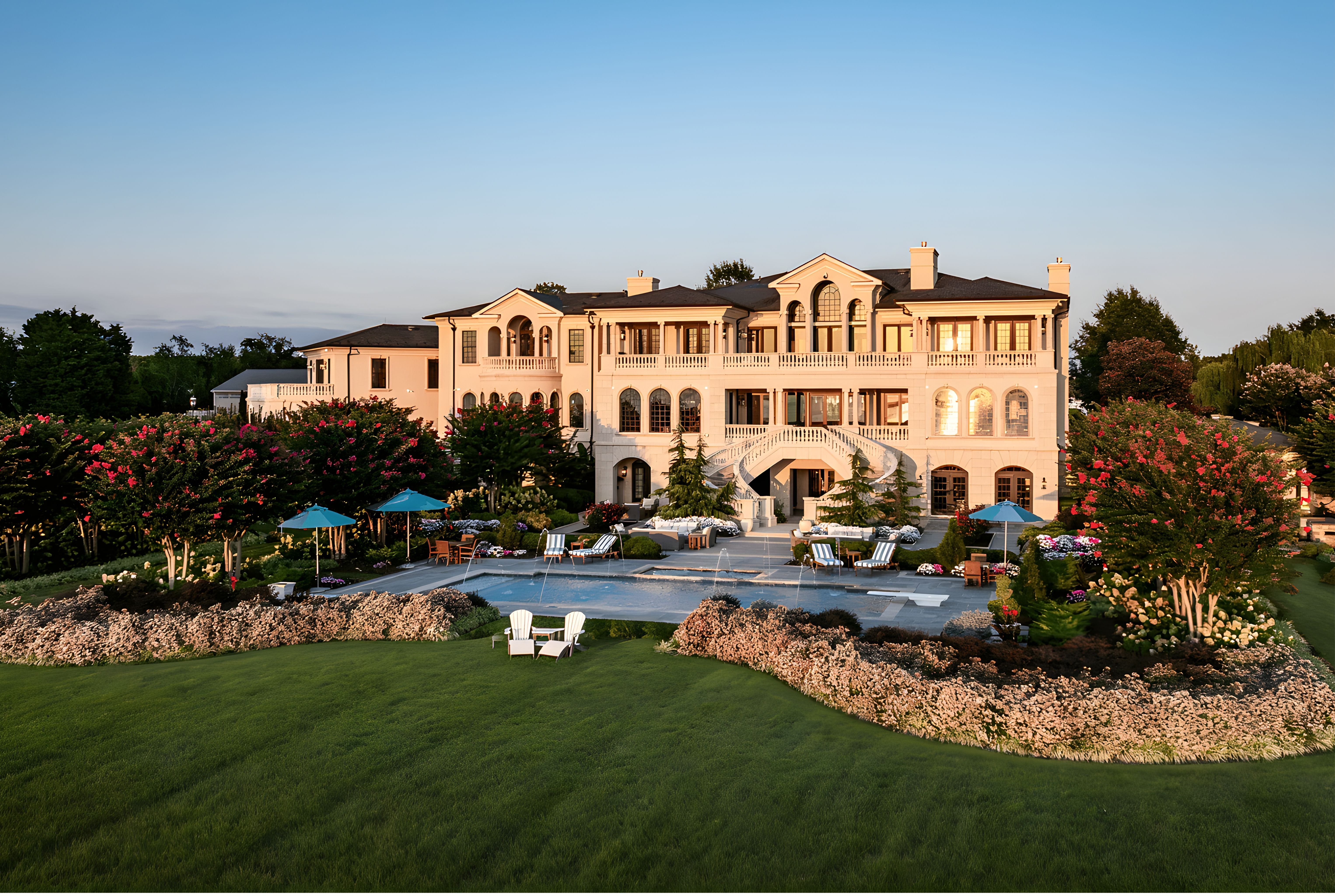 Large beige mansion with multiple windows and curved staircases overlooking a rectangular pool with fountains, surrounded by green lawn, flower beds, and trees with pink blooms under a clear blue sky.